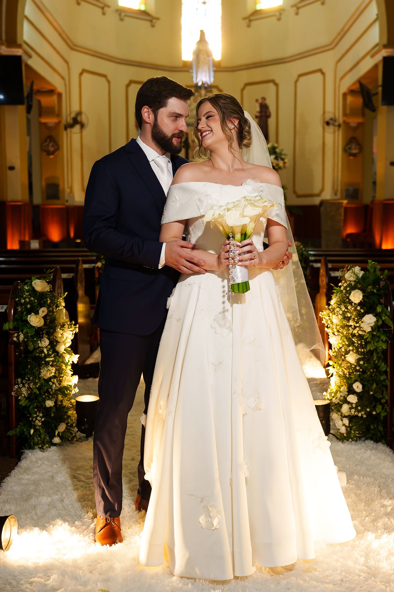 Bride in white off-shoulder gown holds bouquet as groom in navy suit embraces her in a beautifully decorated church aisle.