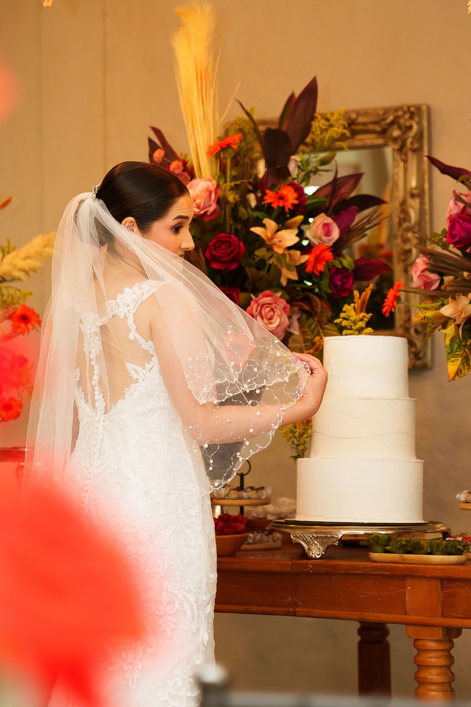 Bride in lace wedding gown and veil cutting a three-tier white wedding cake at a floral-decorated reception table.