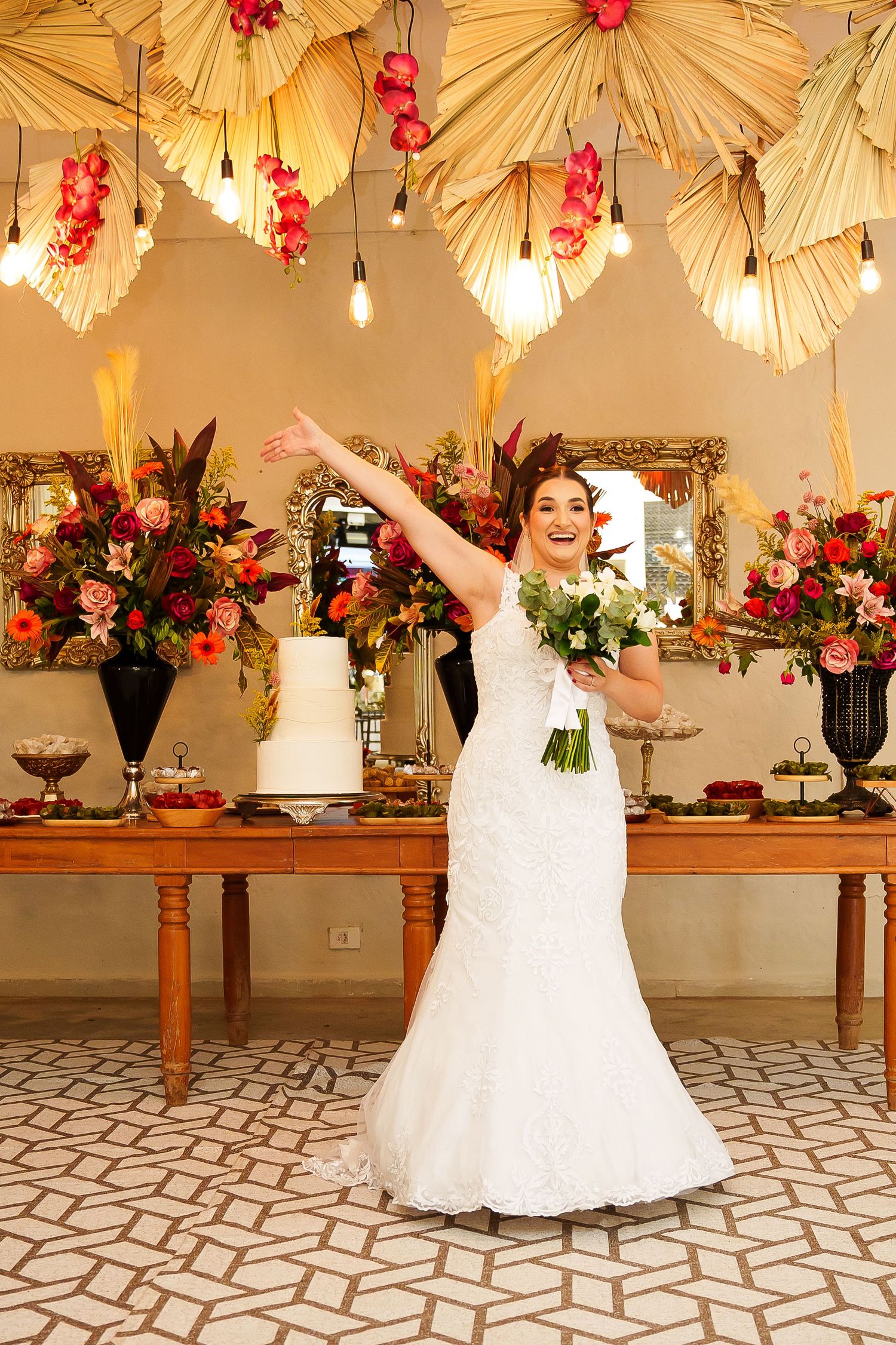 Bride in white lace gown joyfully poses with bouquet at elegant wedding reception with floral decor and cake table.