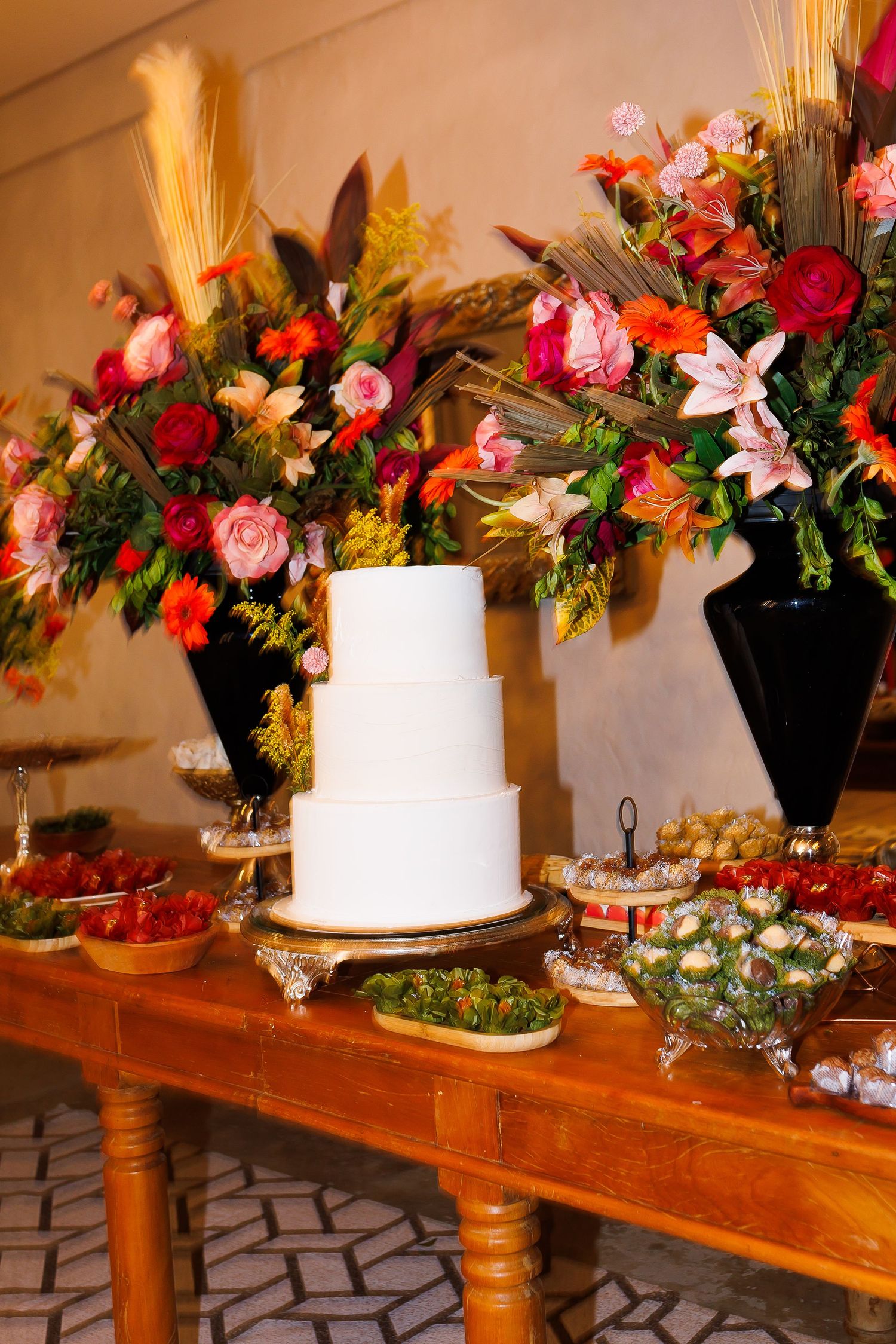 Three-tier white wedding cake on wooden table with colorful floral arrangements and desserts at elegant reception.