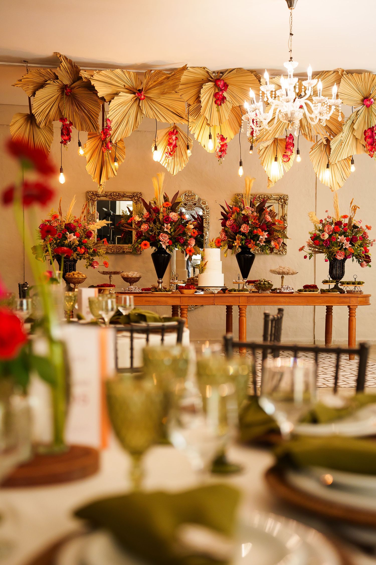 Elegant event decor with gold palm leaf fans, floral arrangements, green glassware, and chandelier lighting.