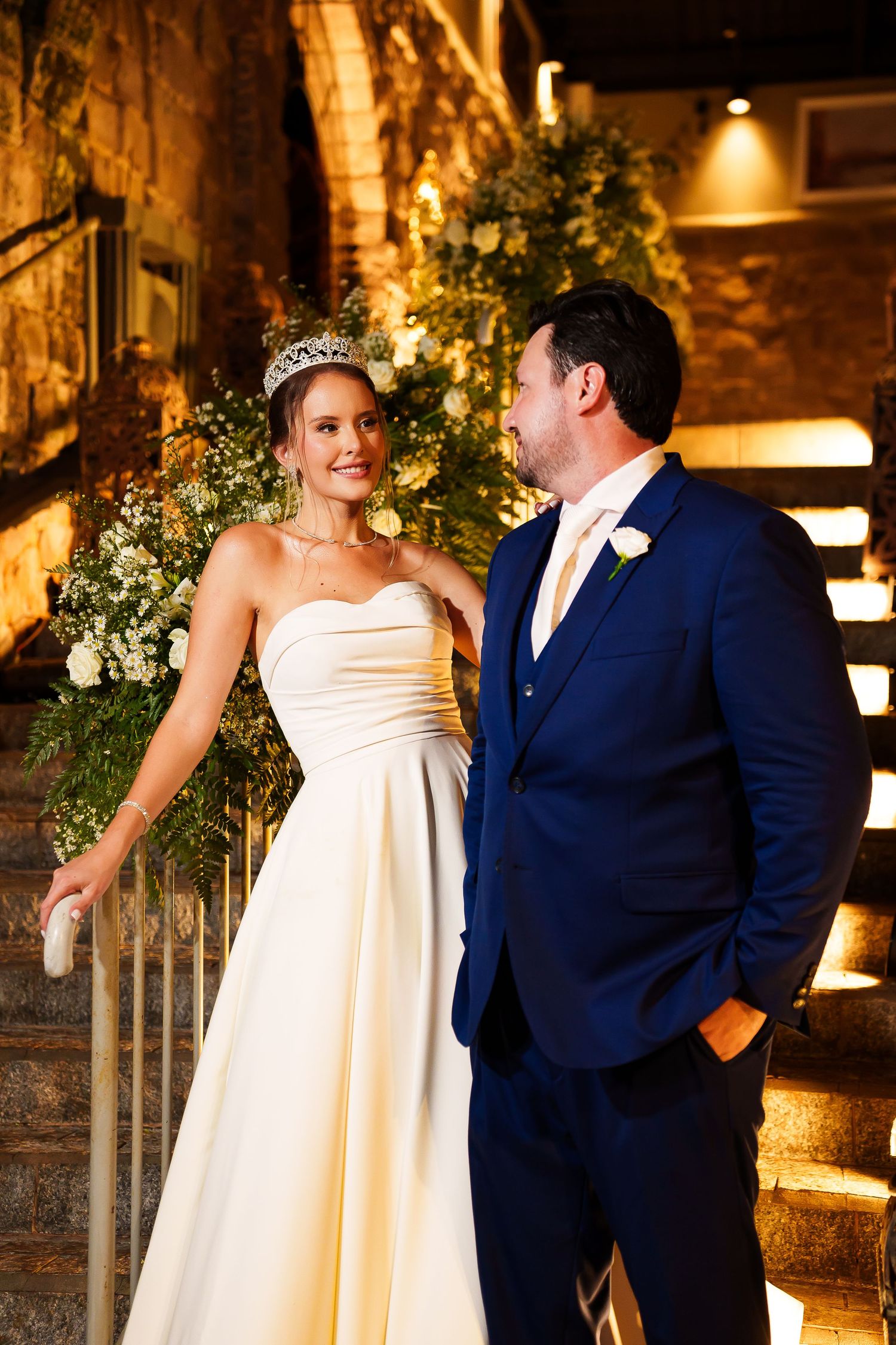 Bride in white gown and tiara smiles at groom in navy suit on elegant staircase with floral backdrop.