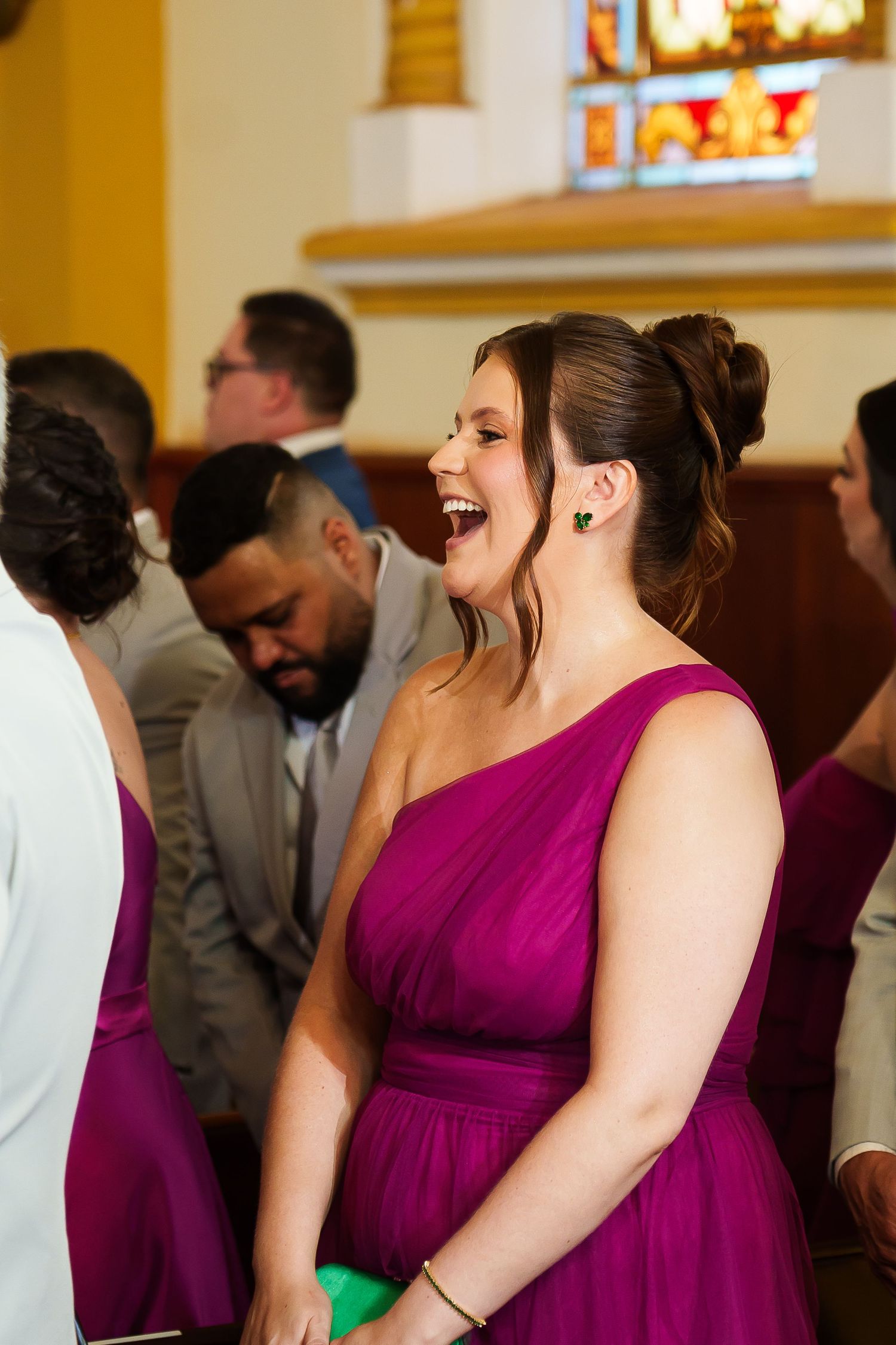 A bridesmaid in a purple one-shoulder gown laughs joyfully inside a church during a wedding ceremony.