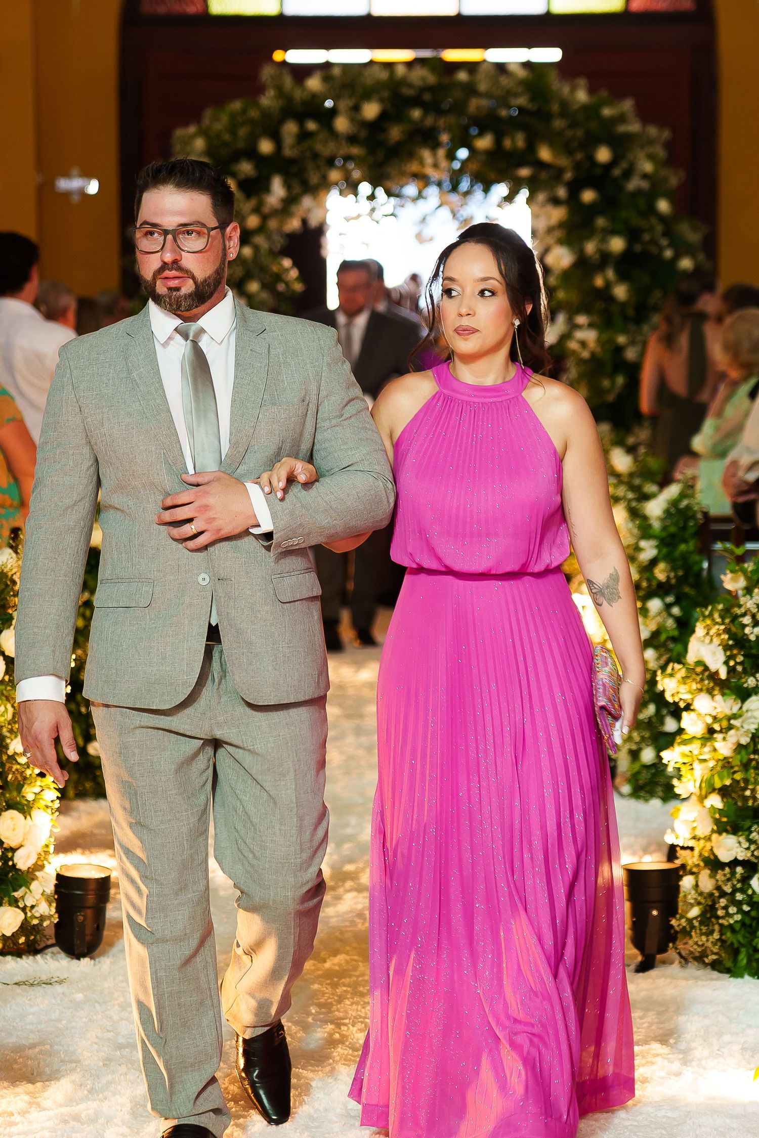Couple walking arm in arm at a wedding, man in gray suit, woman in vibrant pink pleated halter gown, floral arch backdrop.