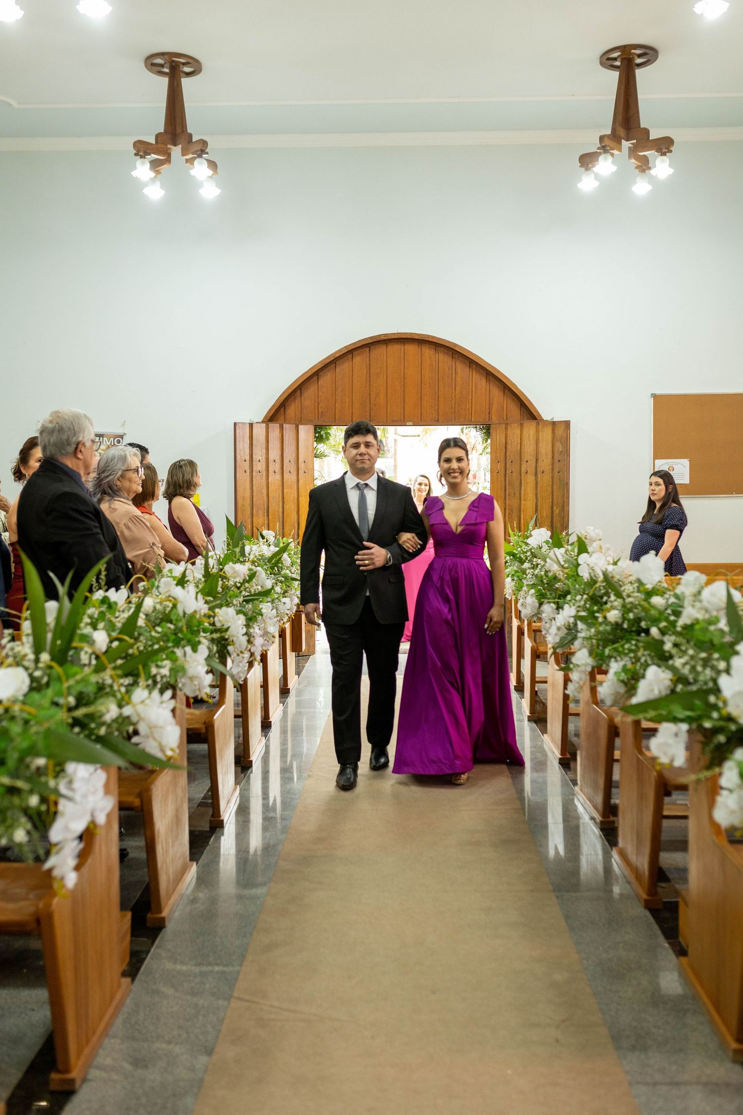 A couple walks down a flower-adorned church aisle, the woman in a purple gown, during a wedding ceremony.