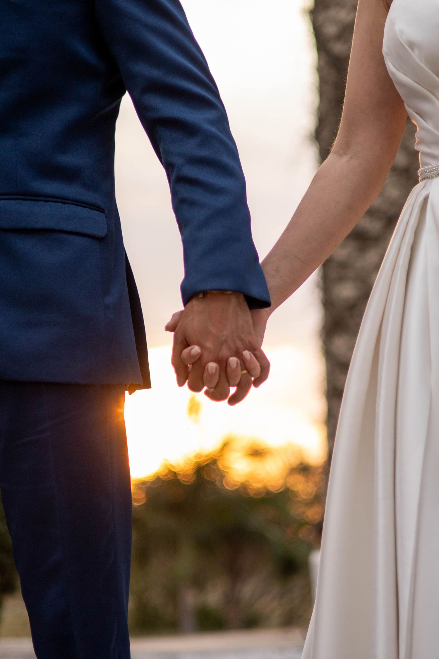 Bride and groom holding hands at sunset, wearing white dress and blue suit, with warm golden light in the background.