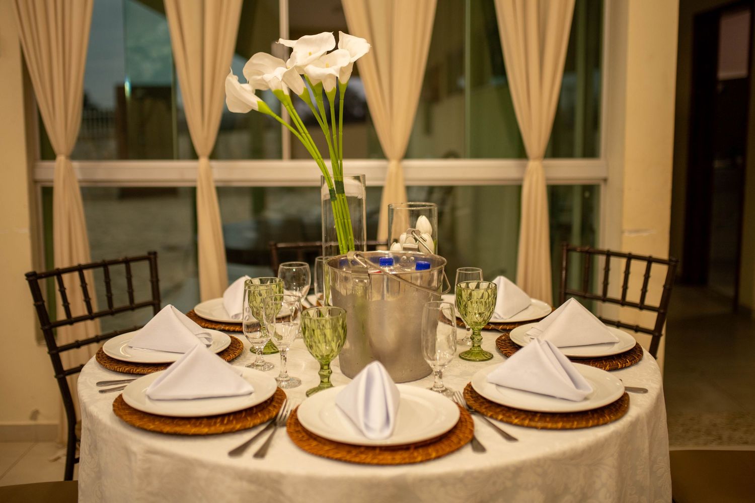 Elegant round dining table set with white linens, green goblets, gold chargers, and white calla lily centerpiece.