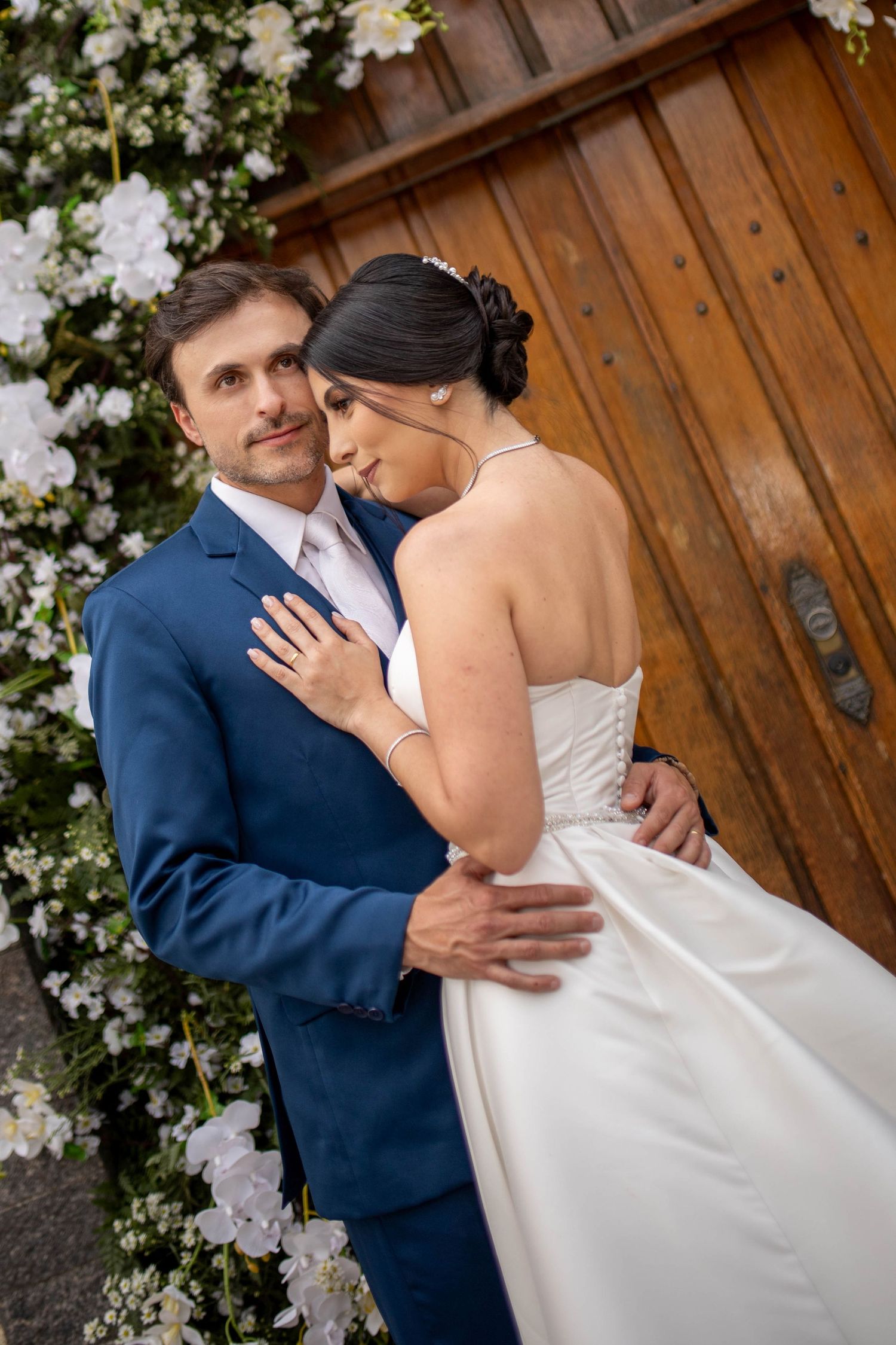 Bride in white gown embraces groom in blue suit before wooden door adorned with white flowers.
