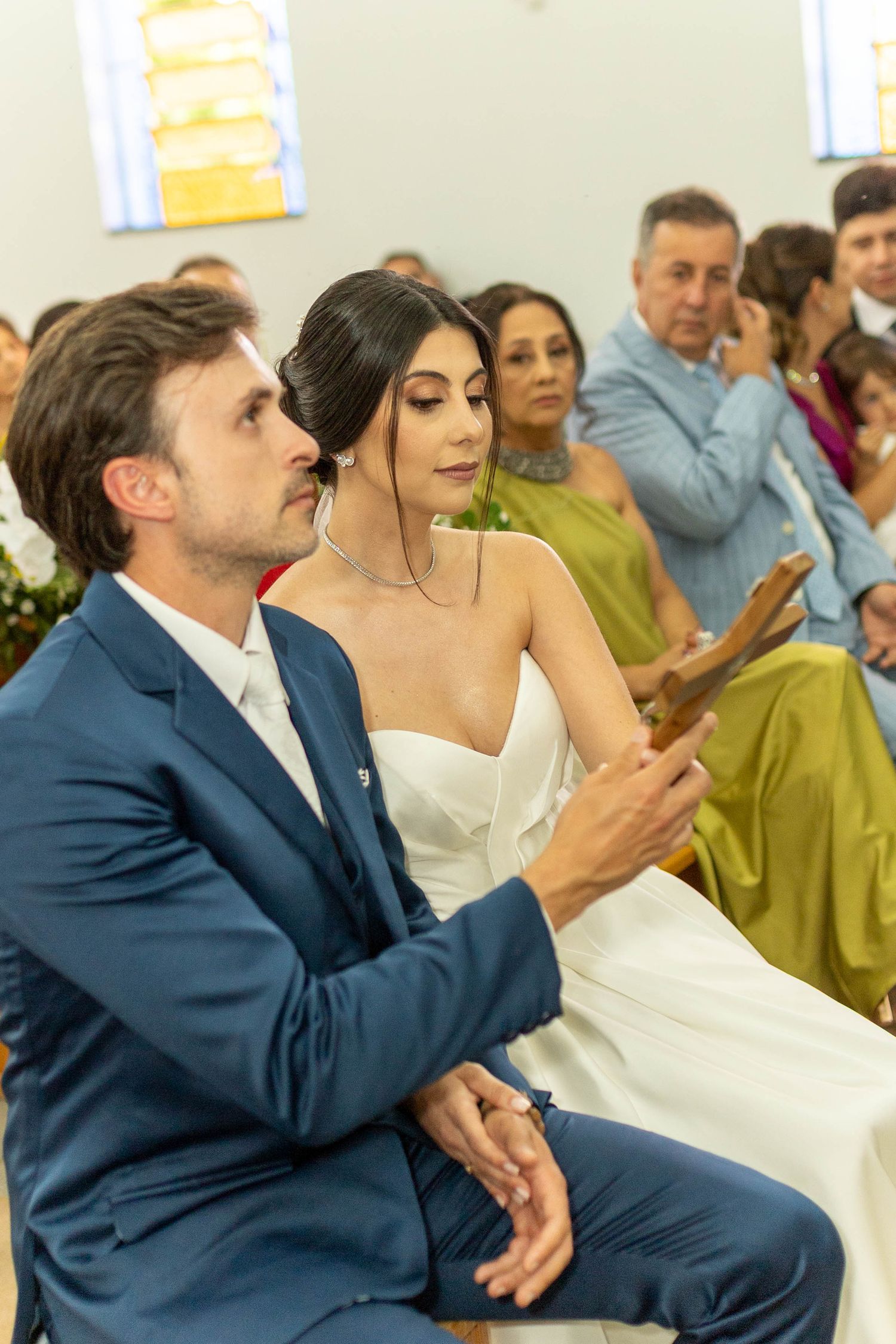 Bride and groom sitting together during wedding ceremony in church, holding wooden cross, guests seated behind them.