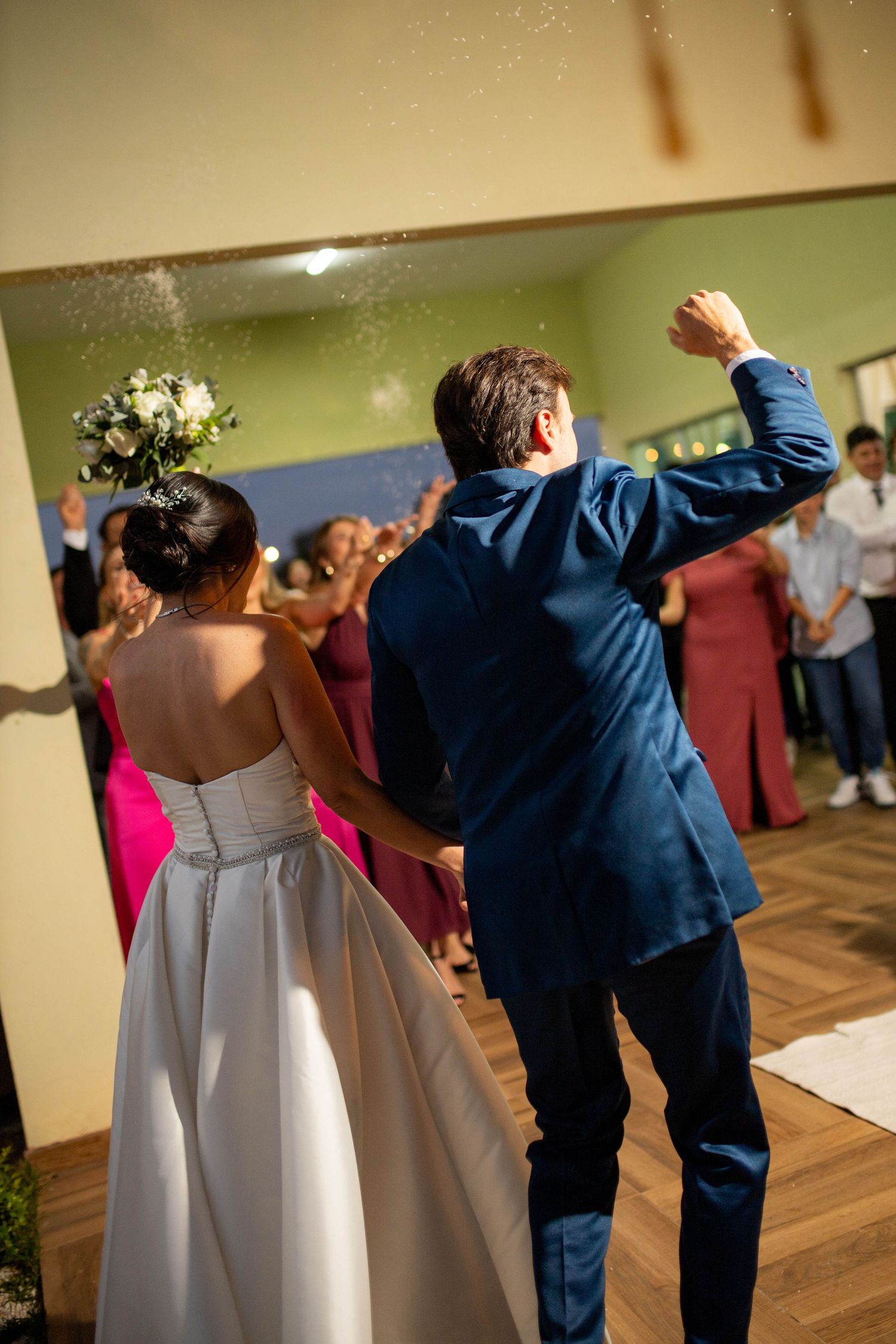 Bride and groom make grand entrance at wedding reception, guests cheering as groom pumps fist in air.