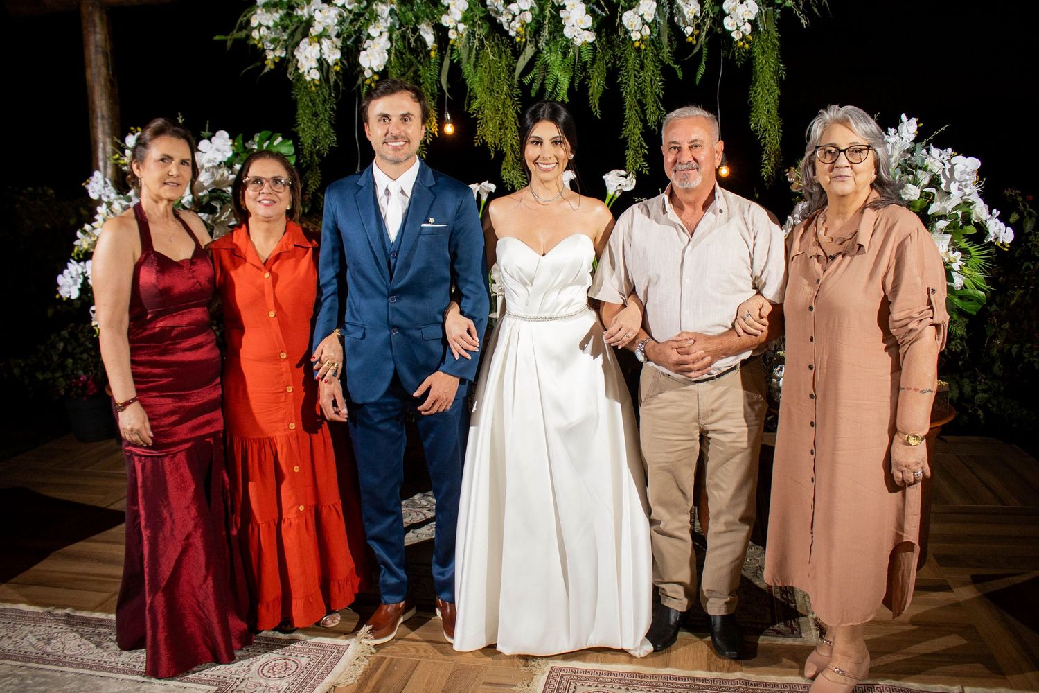 Wedding couple poses with four family members in formal attire under floral arch at elegant outdoor reception.