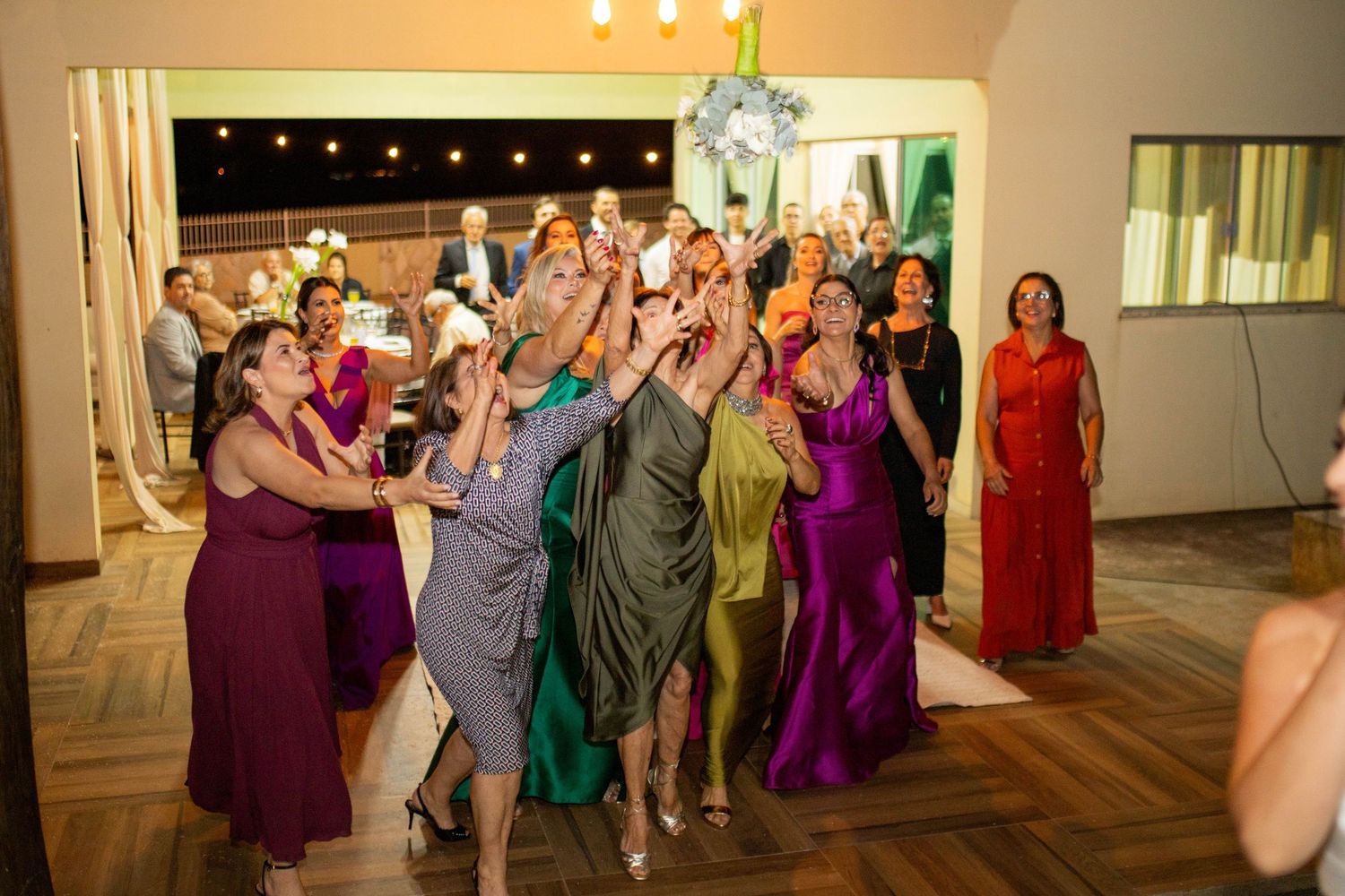 Women in colorful gowns reach up to catch a wedding bouquet toss at an elegant indoor reception venue.