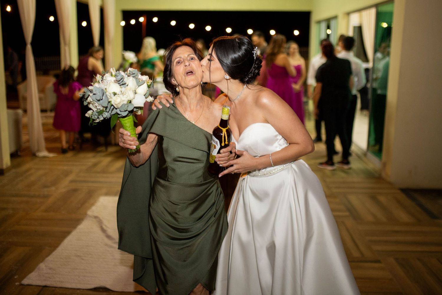 Bride in white gown kisses woman in olive dress holding bouquet at elegant wedding reception with string lights.