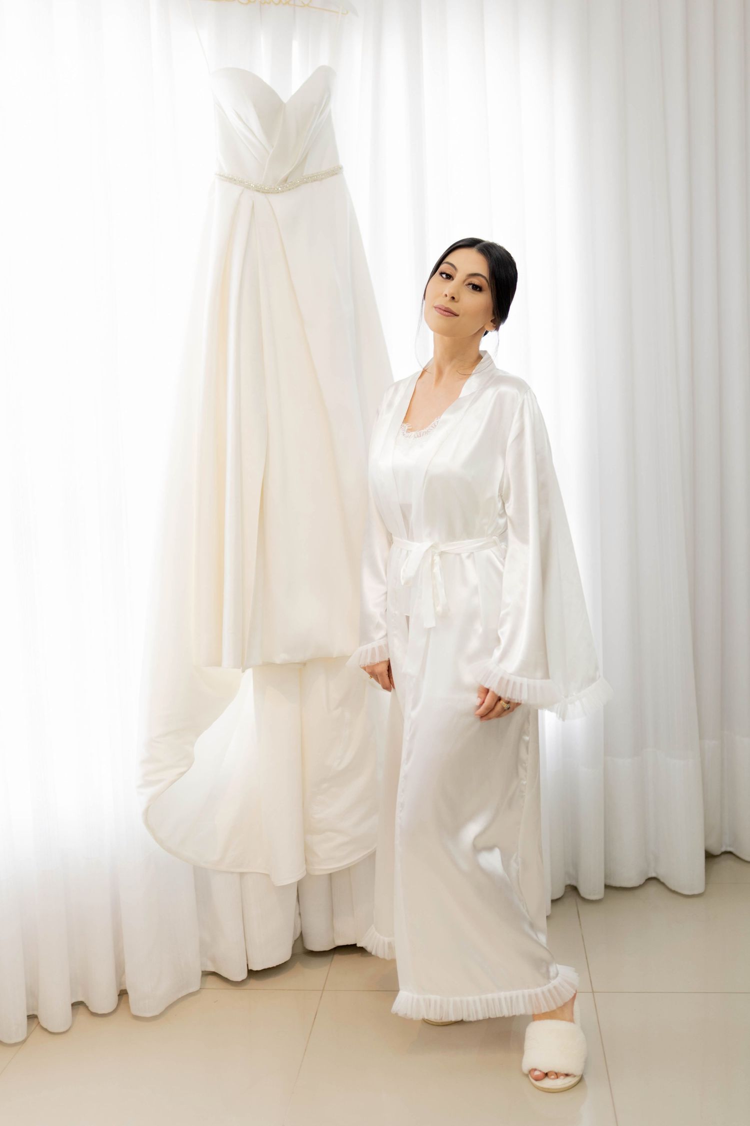 Bride in white satin robe standing next to elegant strapless wedding dress hanging by sheer curtains.