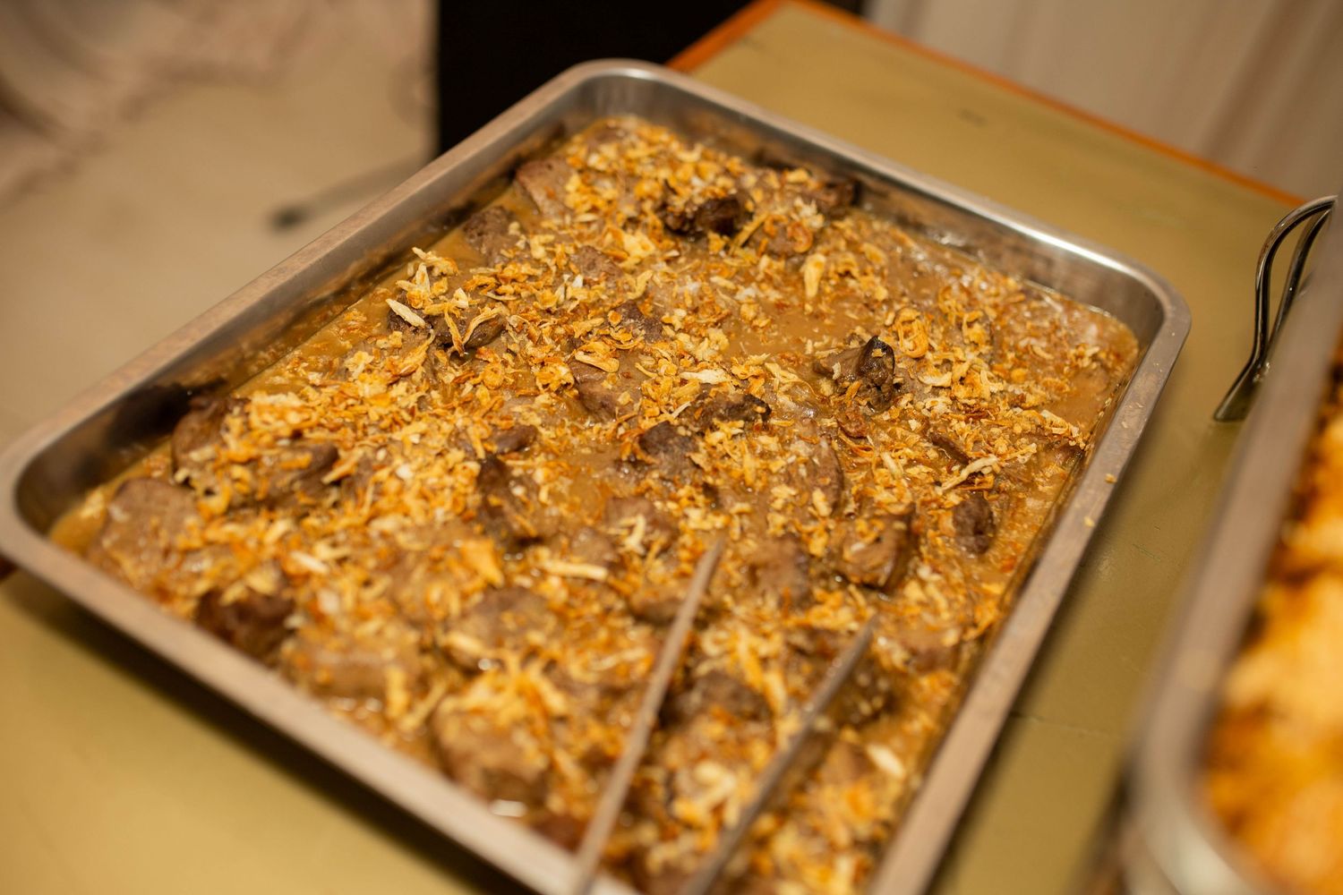 A metal tray filled with seasoned rice and tender meat chunks, garnished with crispy fried onions at a catered event.