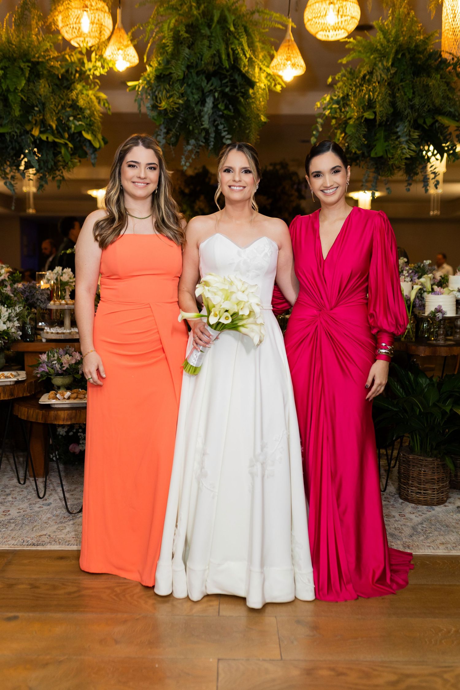 Bride in white gown holds bouquet flanked by bridesmaids in orange and pink dresses at elegant wedding reception.
