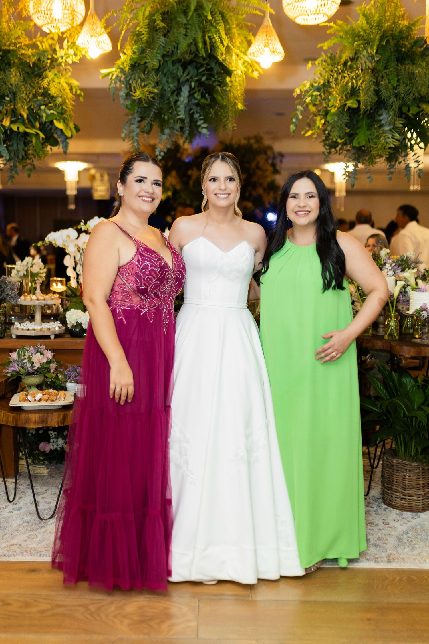 Three women posing together at a wedding reception, bride in white gown flanked by guests in magenta and green dresses.