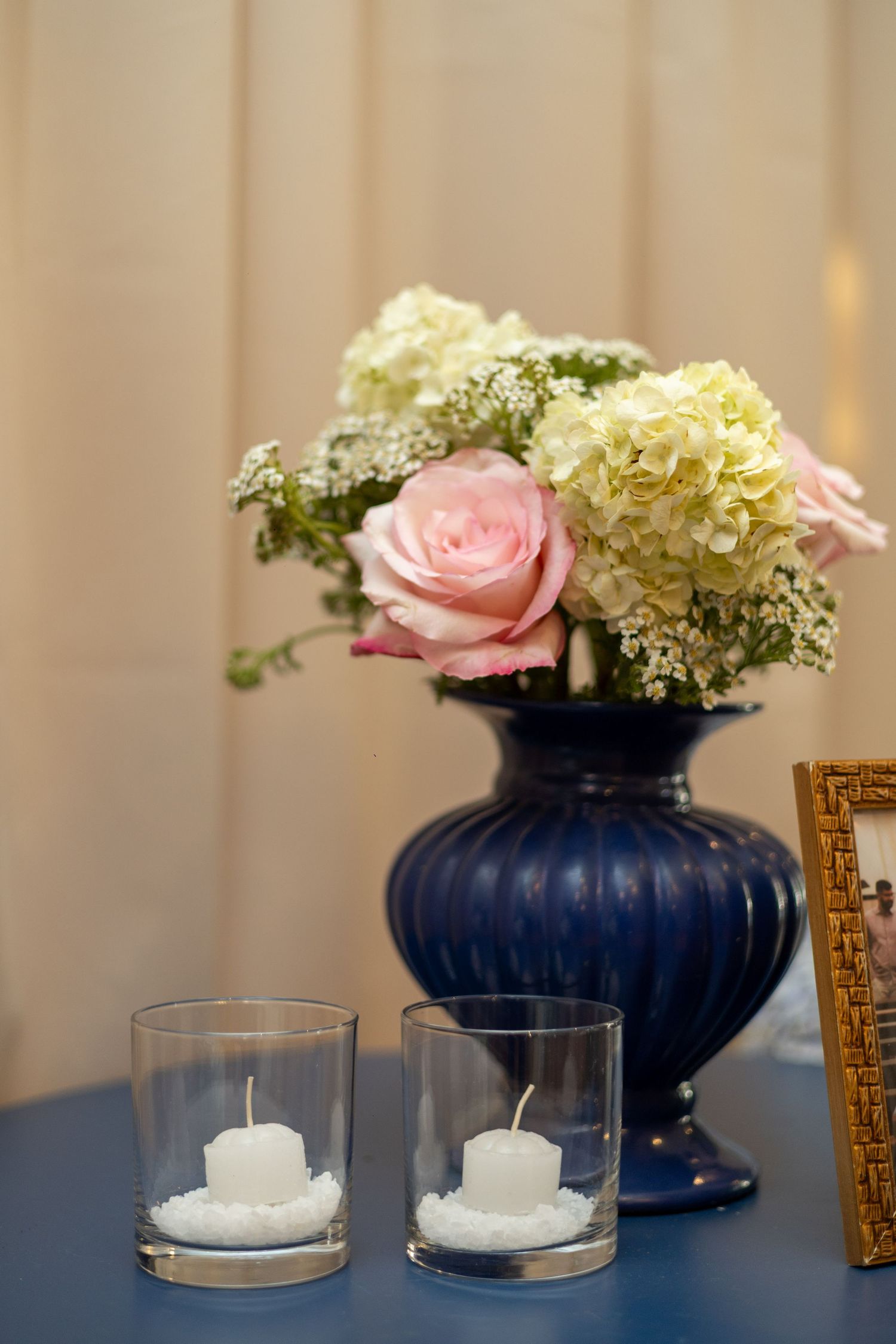 Blue vase with pink roses and white hydrangeas beside two glass candle holders on a blue table.