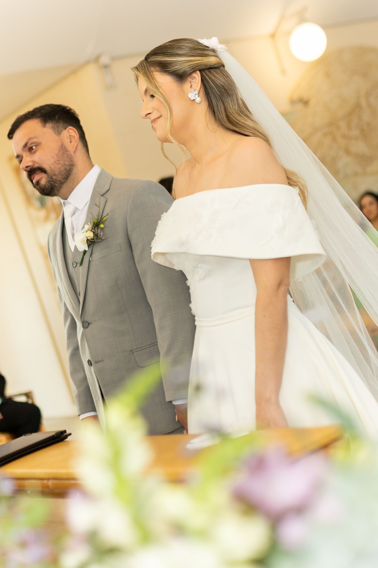 Bride in off-shoulder white gown and veil smiles beside groom in gray suit during wedding ceremony.