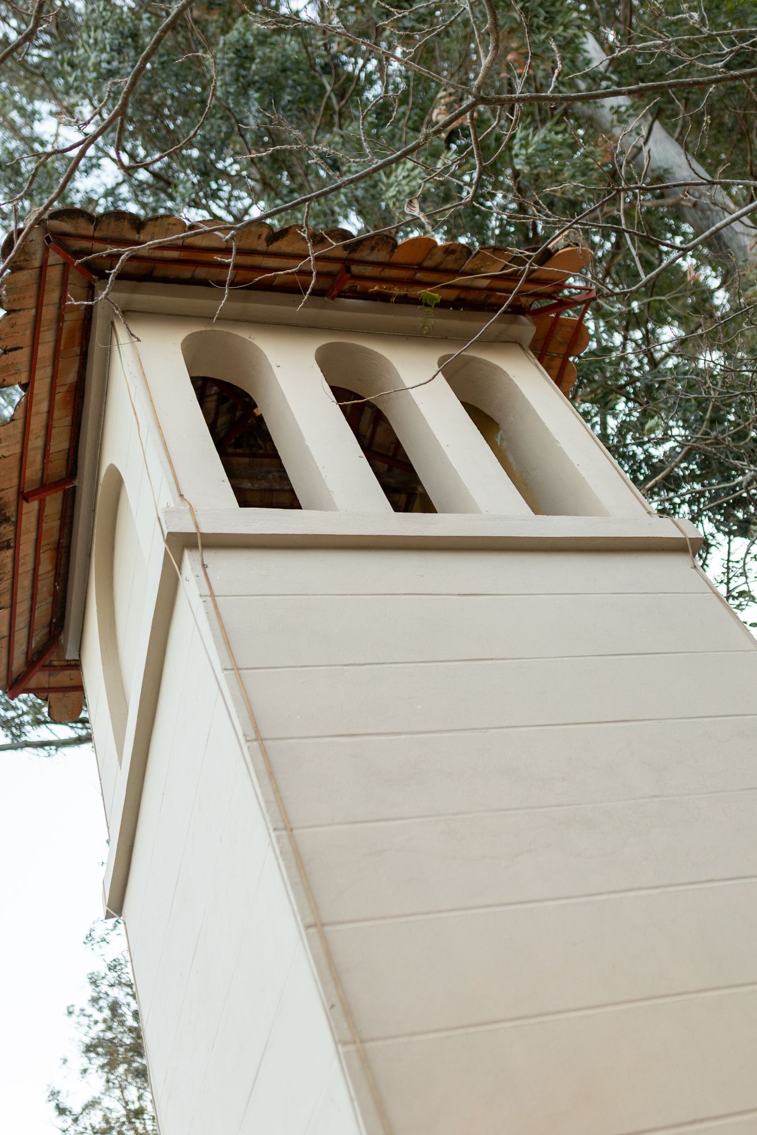 Low angle view of a white church bell tower with arched openings and terracotta roof tiles surrounded by trees.
