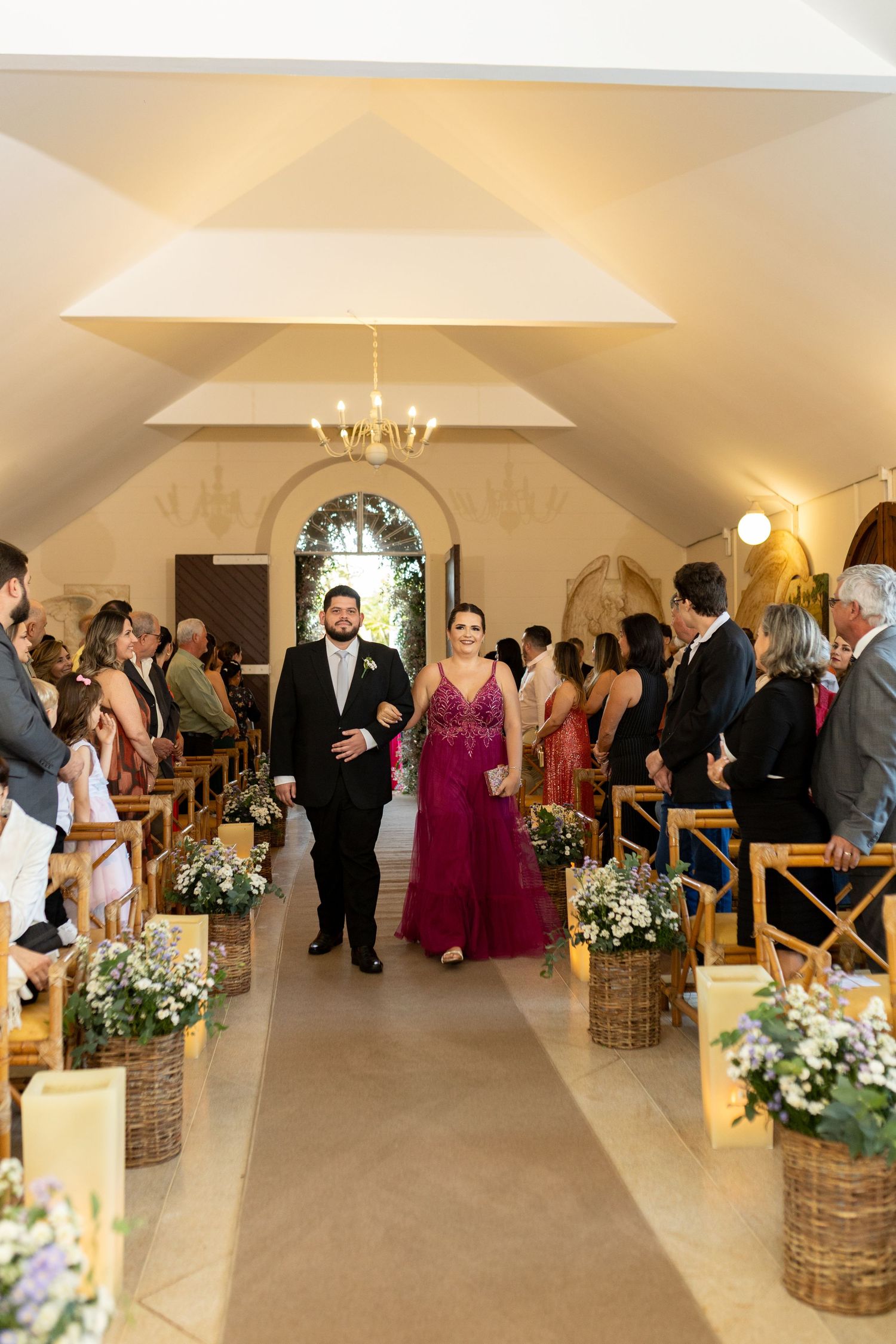 A couple walks down the aisle of an elegant chapel during a wedding ceremony, surrounded by guests and floral arrangements.