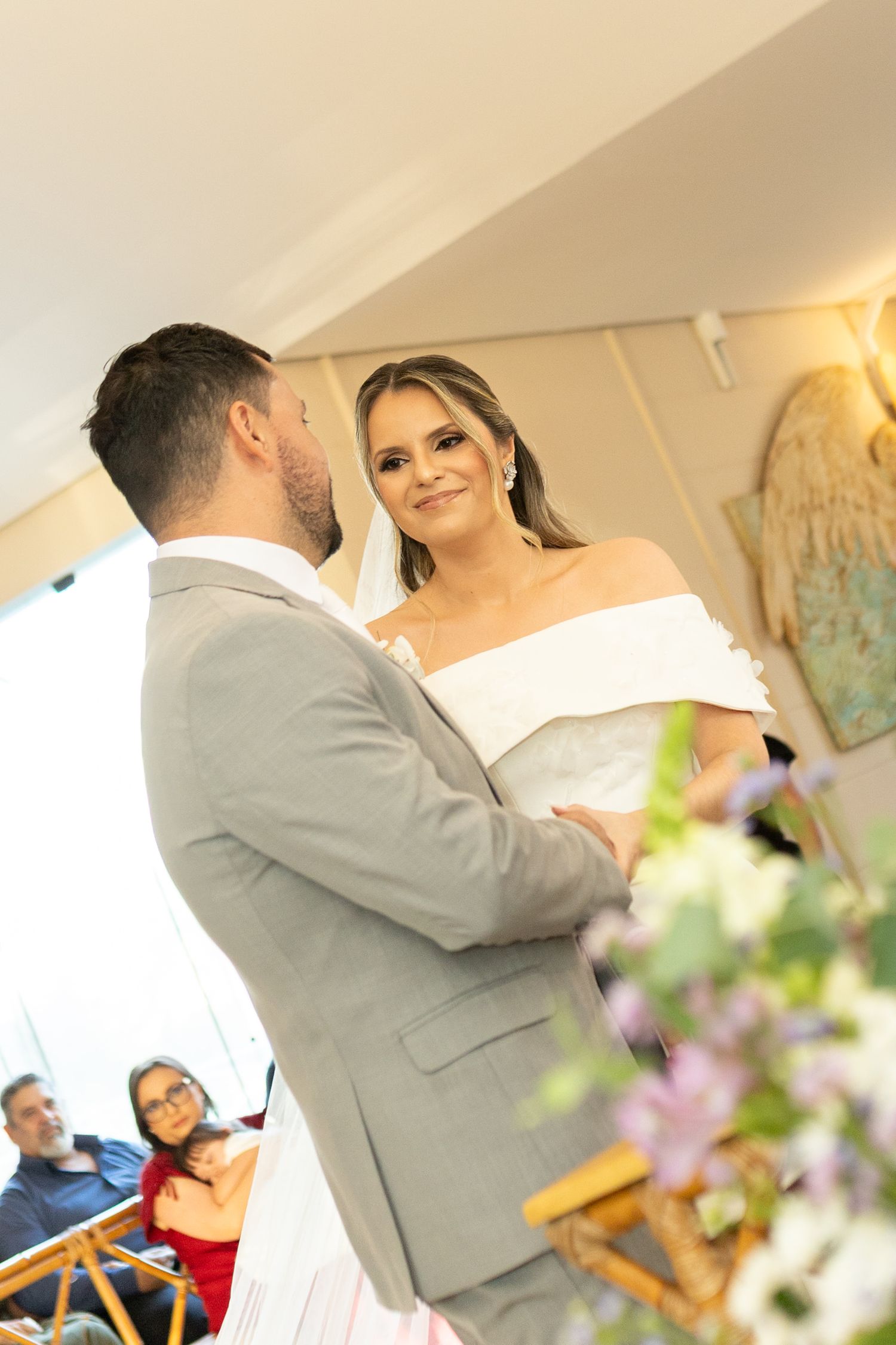 Bride in white off-shoulder gown smiles at groom in gray suit during wedding ceremony with floral decorations.