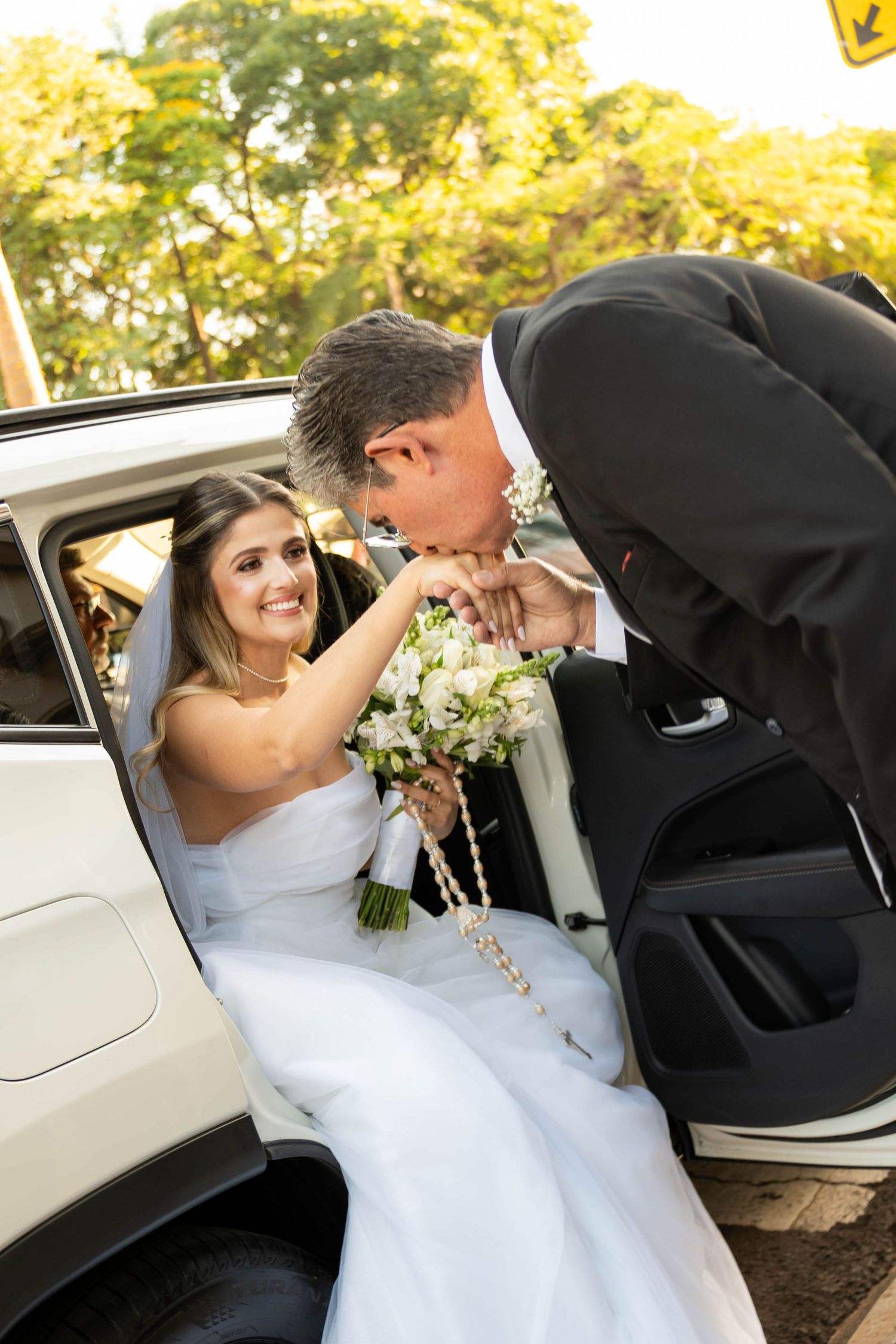 Groom in black suit kissing bride's hand as she sits in car holding white bouquet on wedding day.