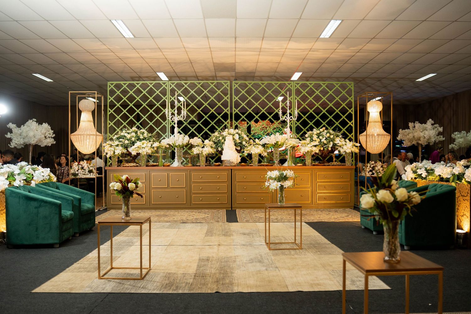 Elegant wedding reception hall with gold decor, floral arrangements, green velvet chairs, and a decorated cake table.