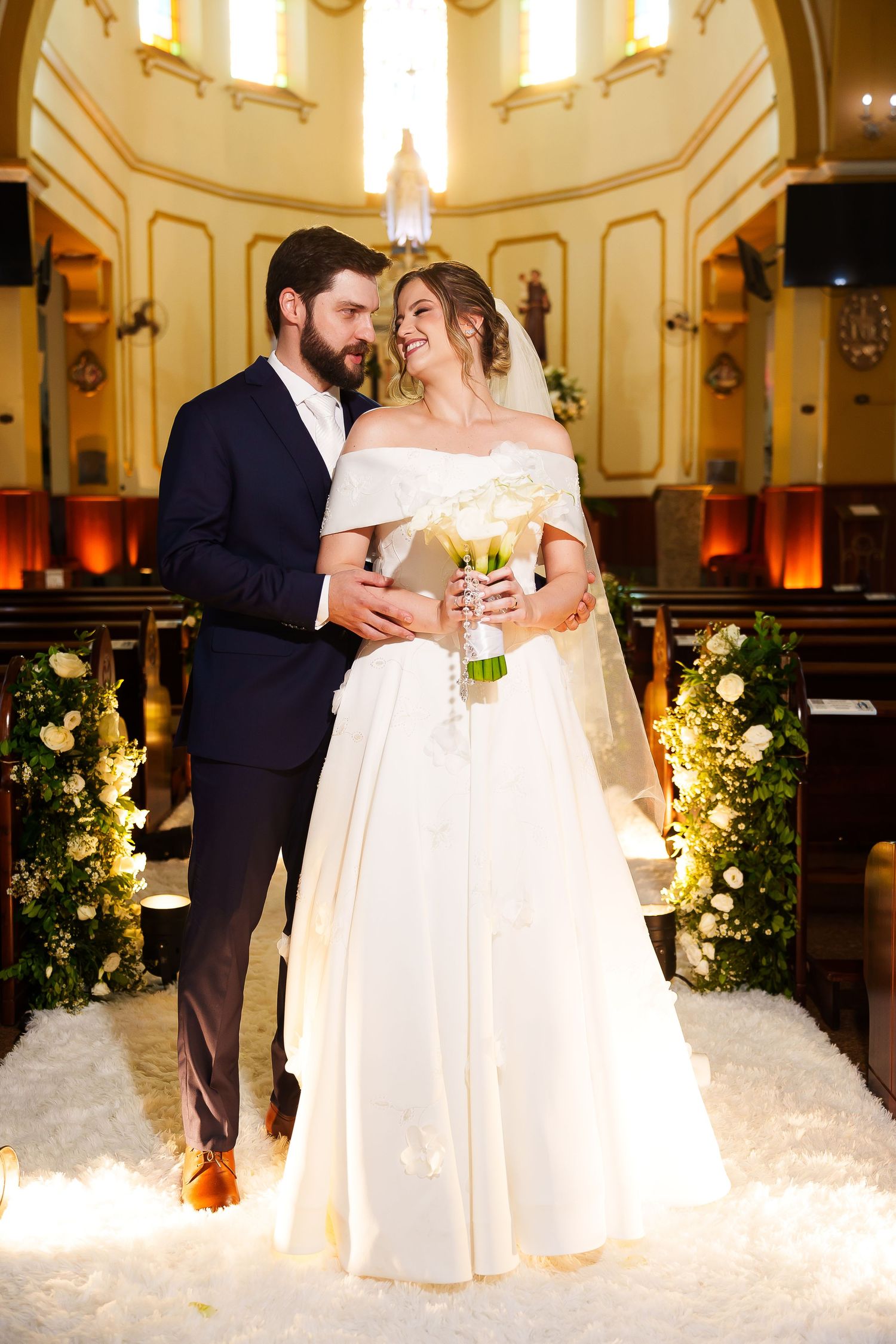 Bride and groom share a romantic moment at church altar, bride in white off-shoulder gown holding bouquet.