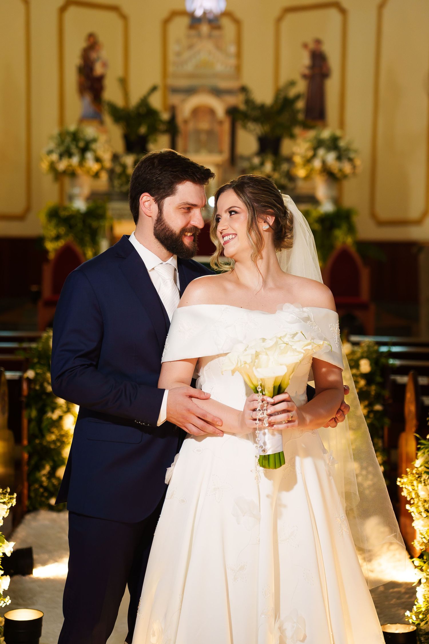 Bride and groom share a romantic moment at church altar, bride holding white bouquet in elegant off-shoulder gown.