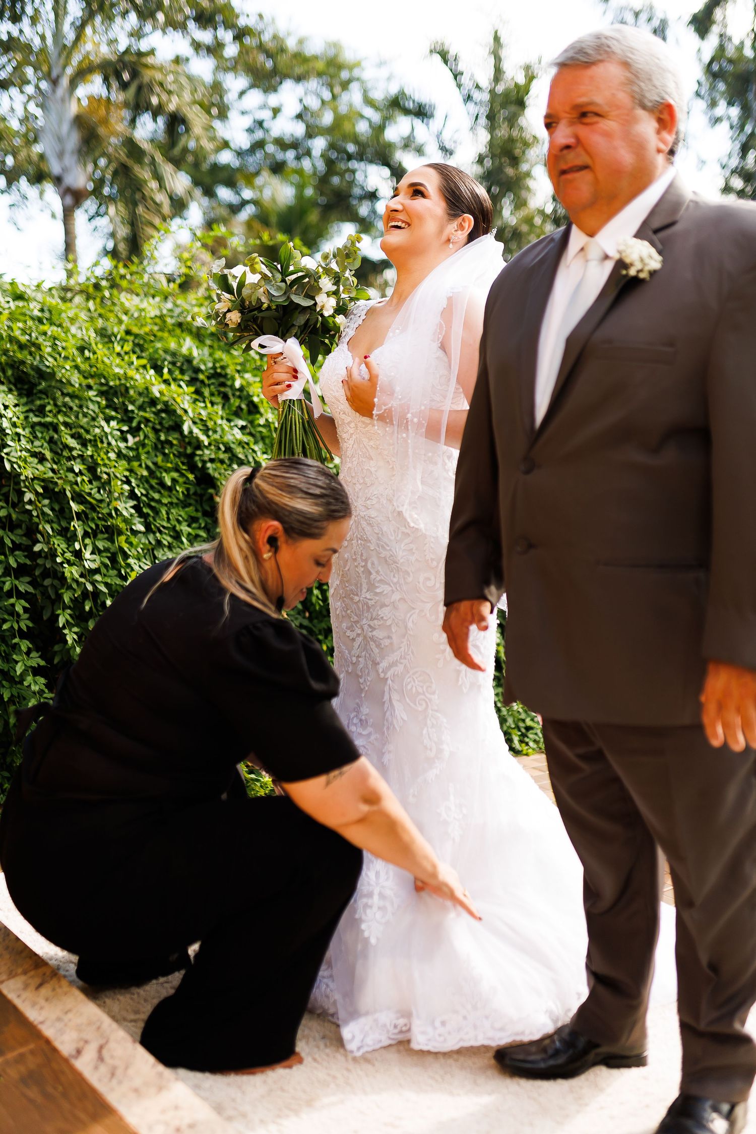 A bride in white lace gown holds bouquet beside suited man as assistant adjusts her dress train outdoors.