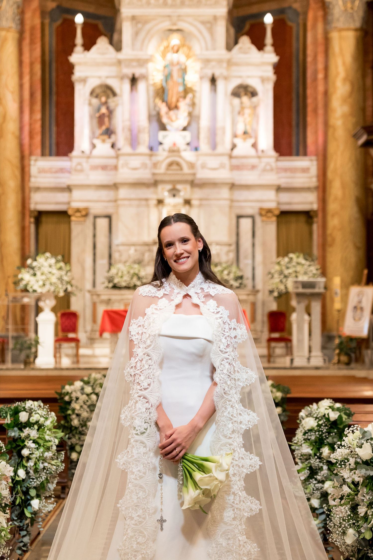 Bride in white gown with lace cape holds calla lily bouquet, standing before ornate church altar.