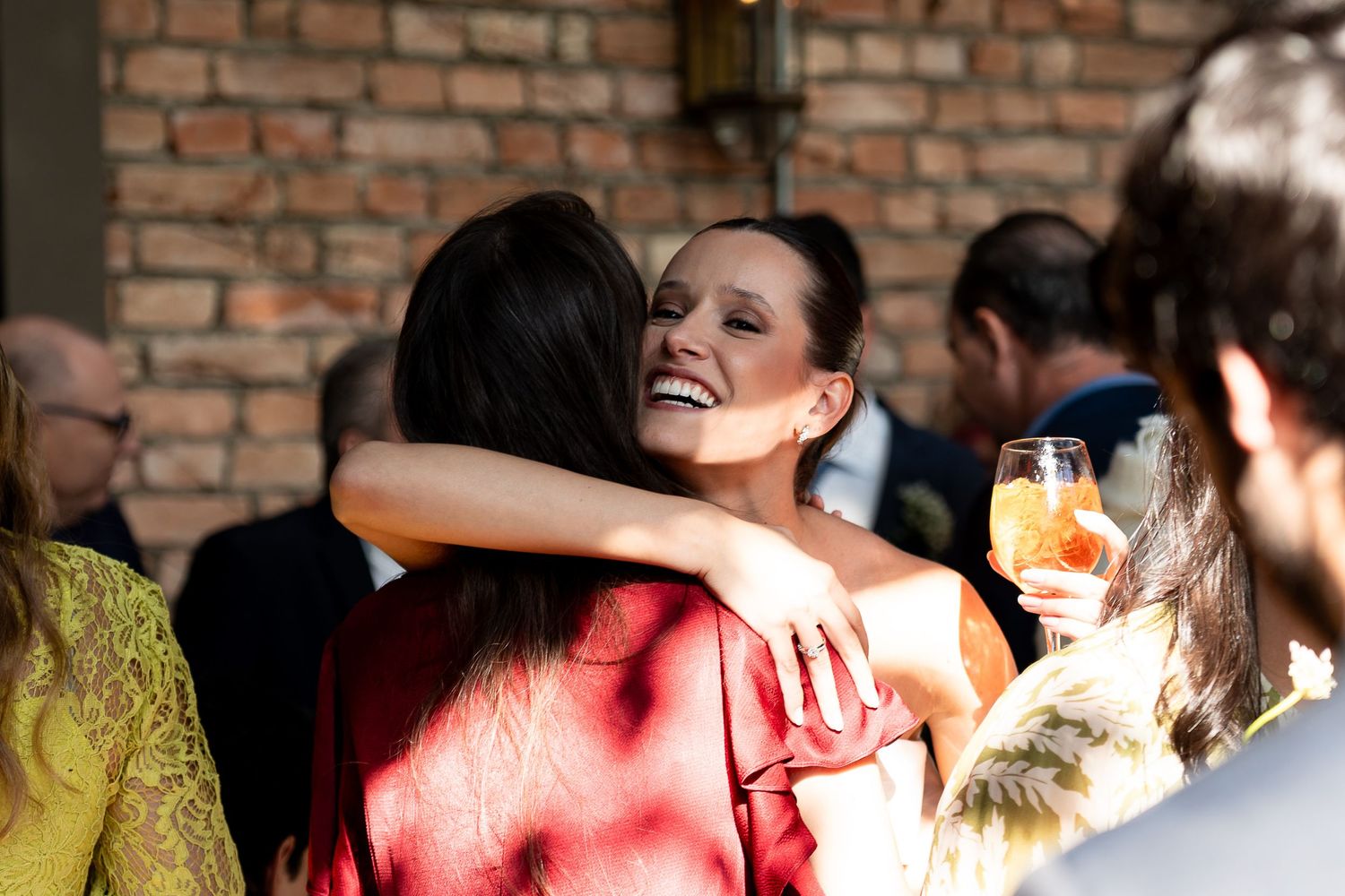 Two women sharing a warm hug at a wedding reception, one smiling joyfully while holding a drink outdoors.