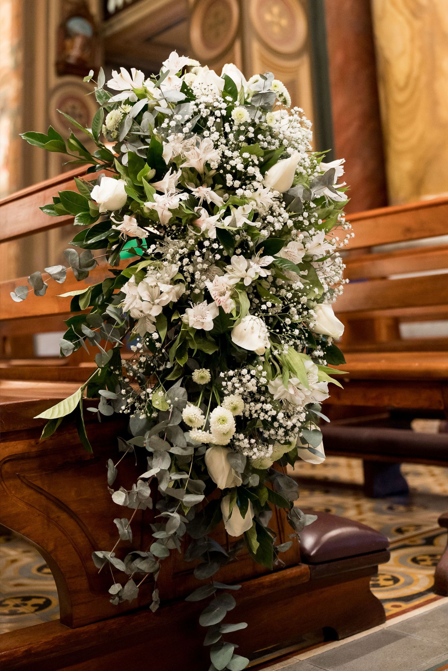Elegant cascading white wedding bouquet with roses, baby's breath, and eucalyptus displayed on a wooden church pew.