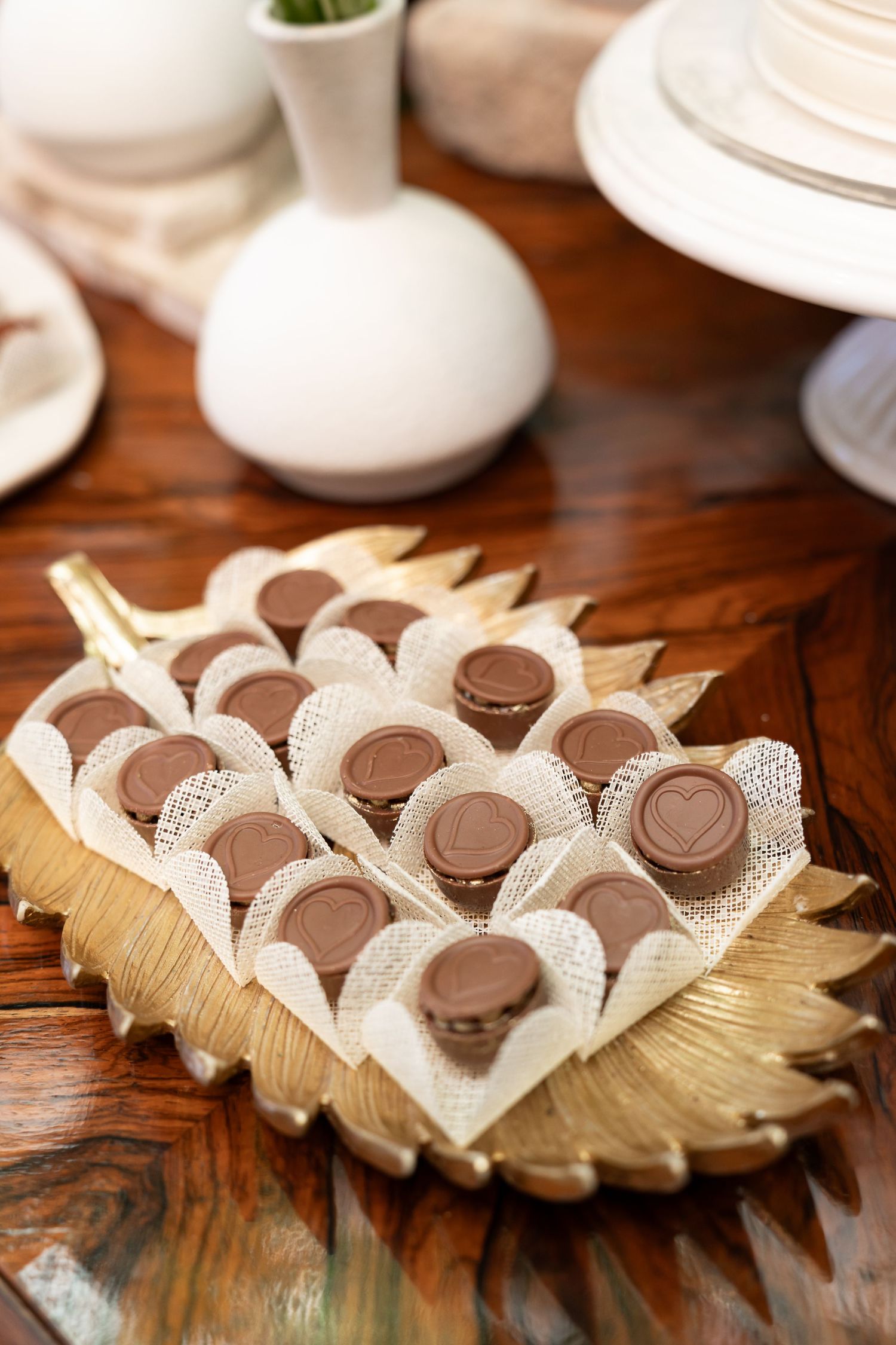 Gold leaf tray displaying chocolate bonbons in lace wrappers on a wooden table with white vases.