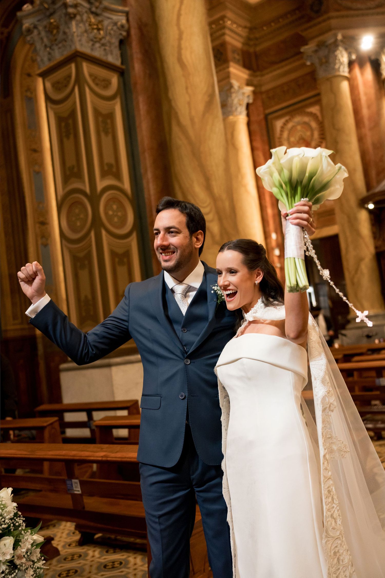 Joyful newlyweds celebrate in ornate church, bride raising bouquet of white calla lilies, groom pumping fist in blue suit.