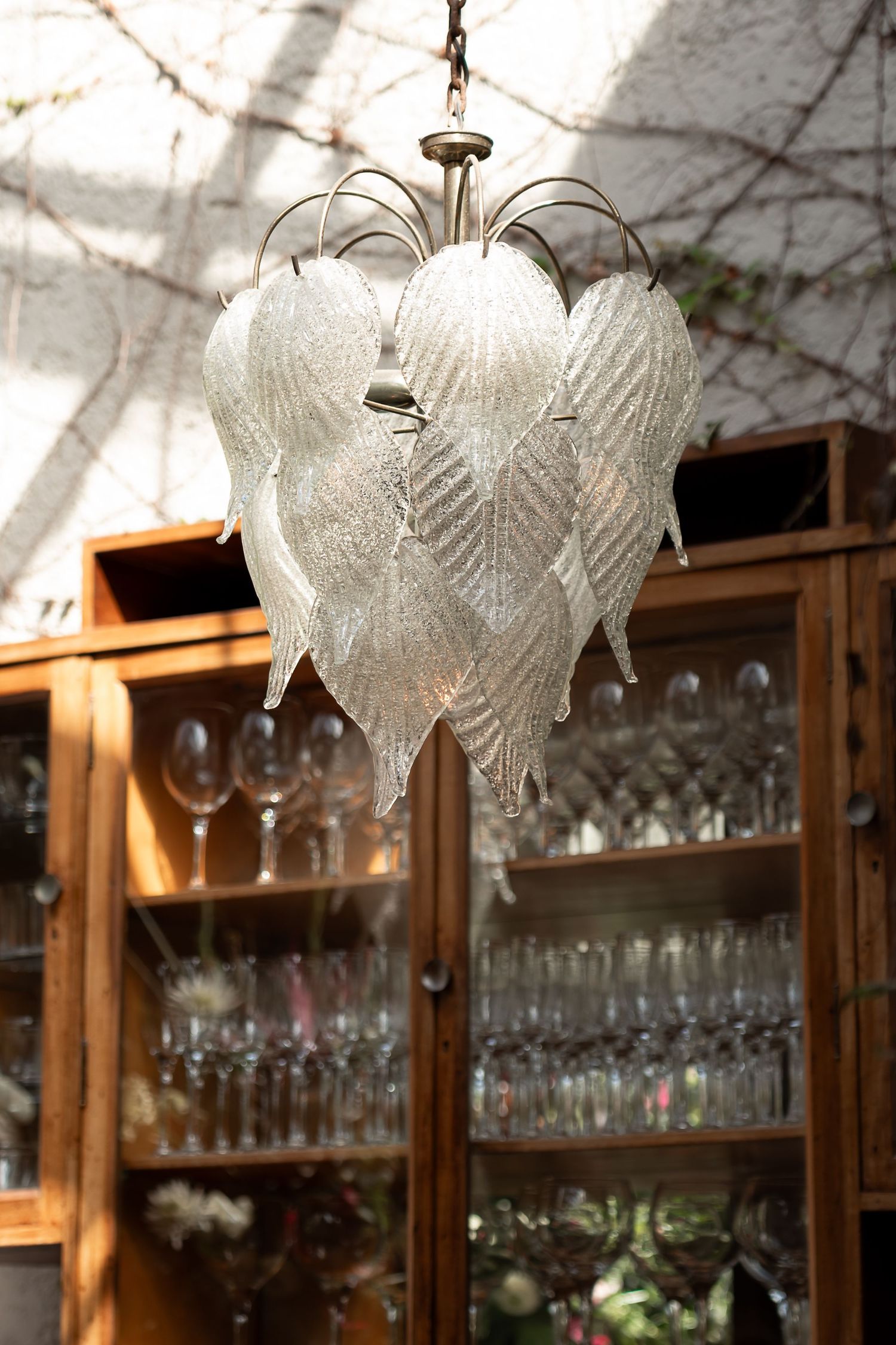 Elegant glass leaf chandelier hanging in front of wooden display cabinets filled with crystal glassware.