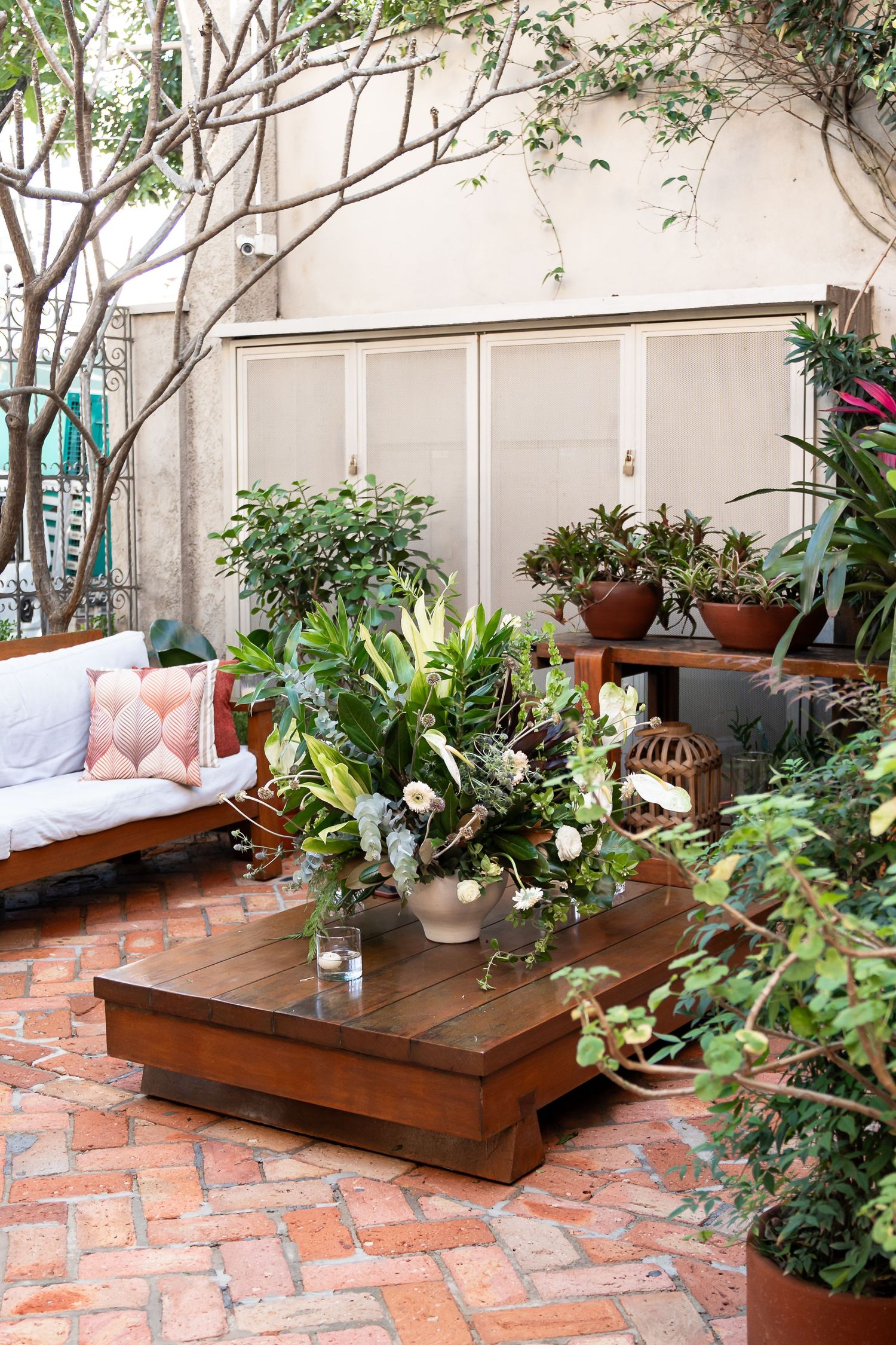 Cozy outdoor patio with brick flooring, wooden coffee table, lush plants, and a white sofa with pink pillows.