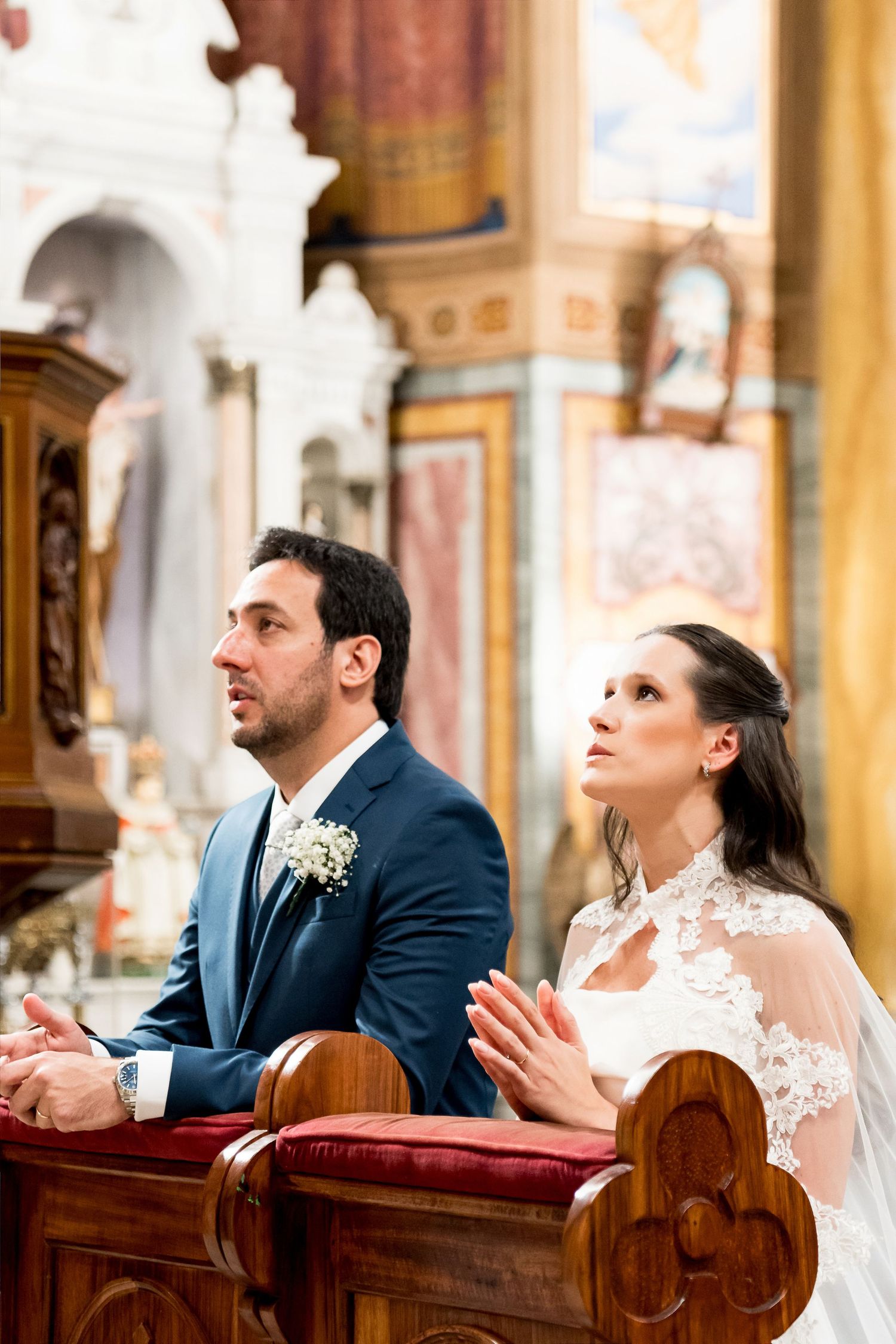 Bride and groom kneeling in prayer at church altar during wedding ceremony, dressed in white gown and navy suit.