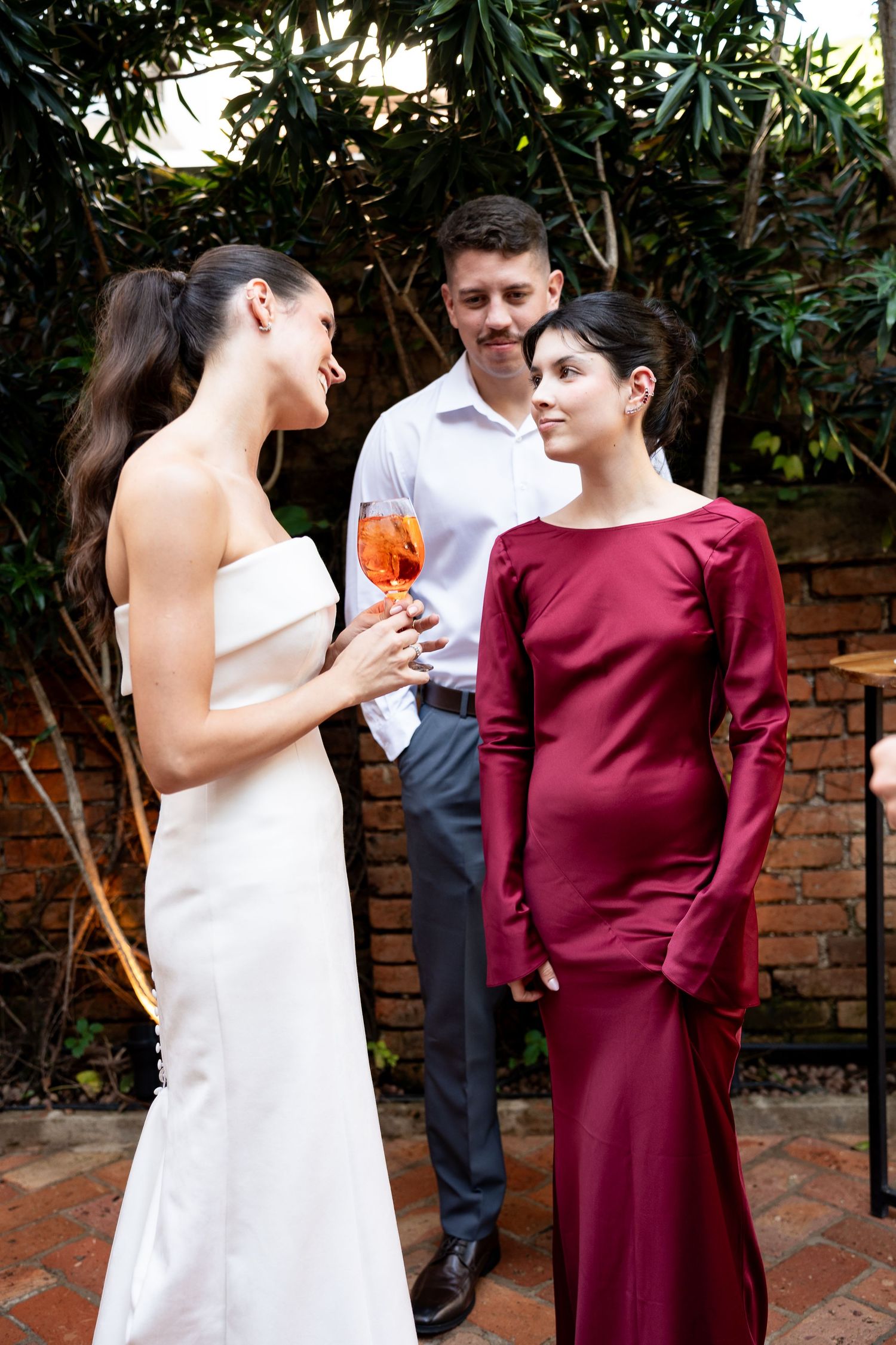 Two women in elegant gowns chat at an outdoor garden party, while a man in a white shirt stands behind them.