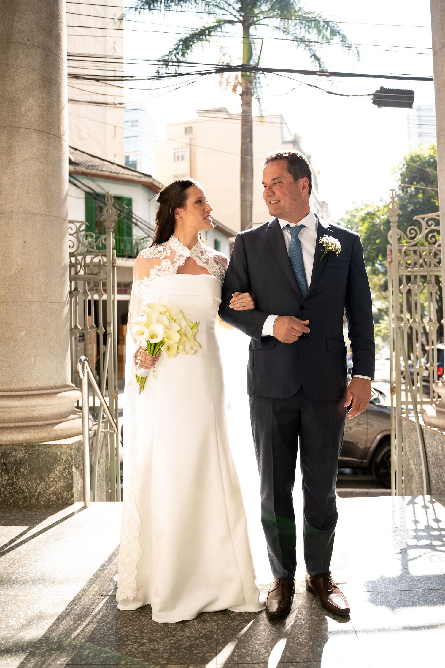 Bride in white lace gown holding calla lilies bouquet stands with groom in navy suit outside church.