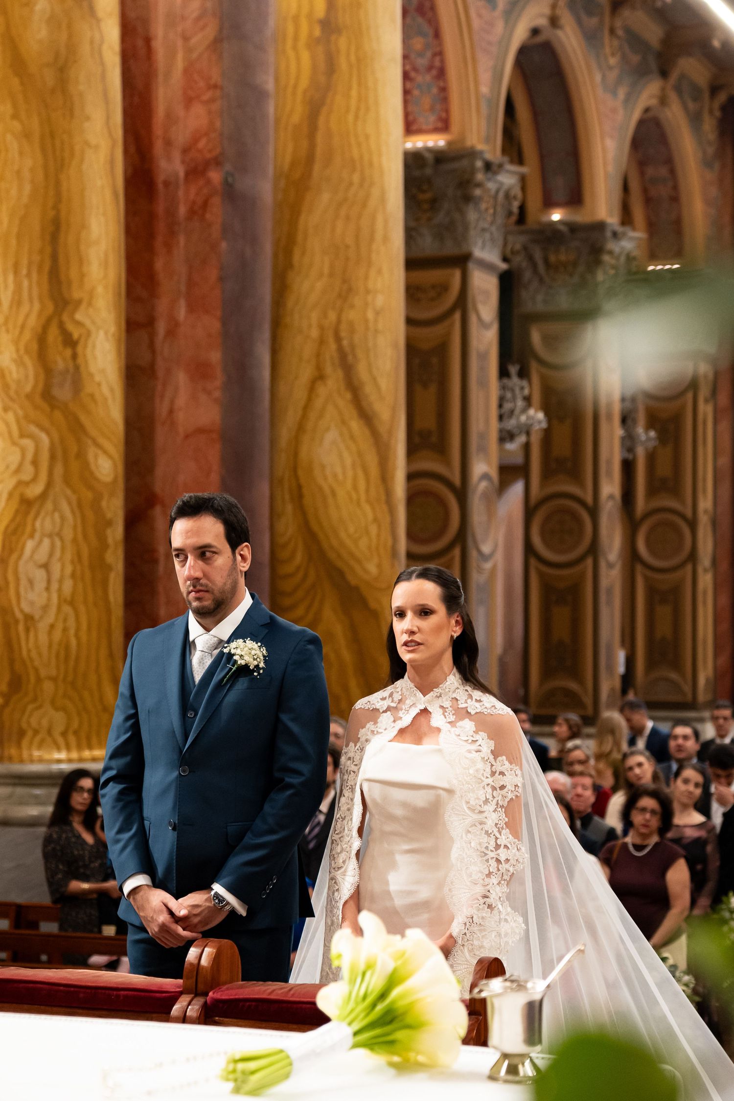 Bride in white gown with lace cape and groom in blue suit stand at church altar during elegant wedding ceremony.