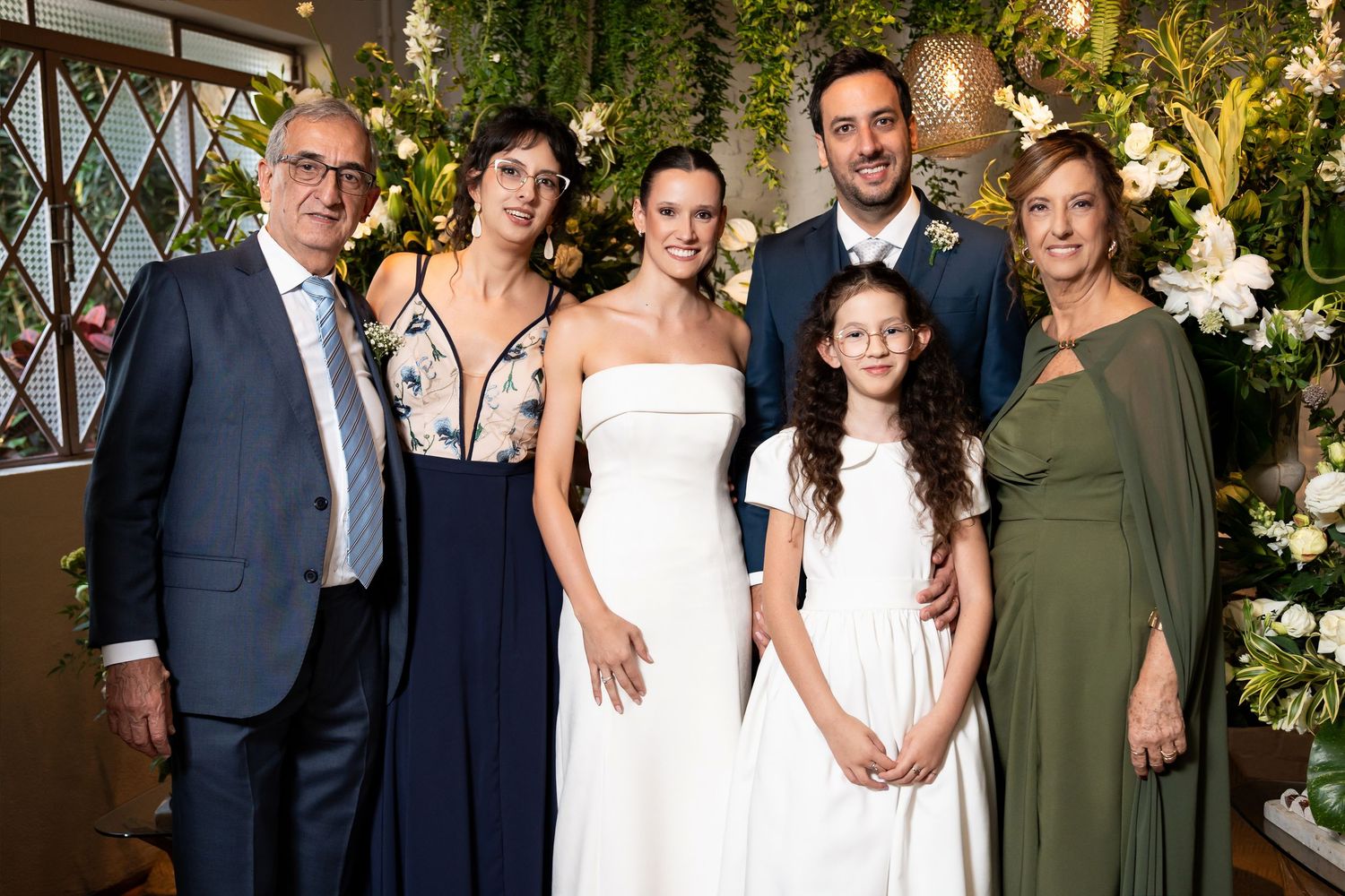 Family group photo at a wedding with bride in white gown, groom in suit, and relatives posing near floral decorations.