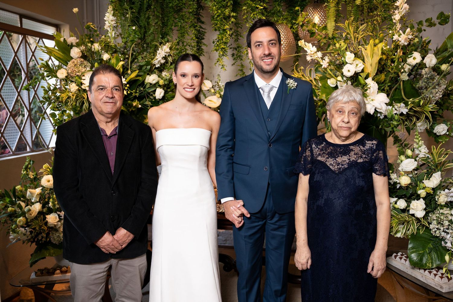 Wedding couple poses with family members in front of lush floral backdrop at elegant indoor ceremony.