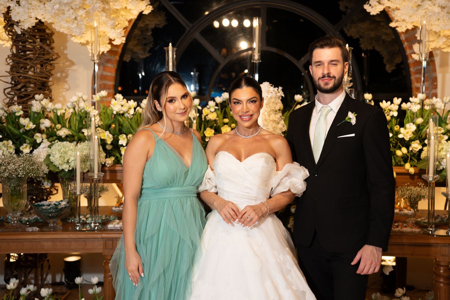 Bride in white gown poses with bridesmaid in teal dress and groom in black suit at elegant floral wedding reception.
