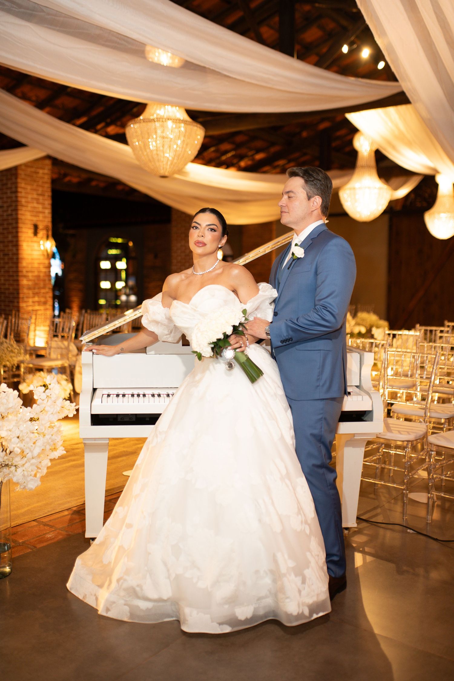 Bride in white ball gown and groom in blue suit pose elegantly at indoor wedding venue with draped ceilings.