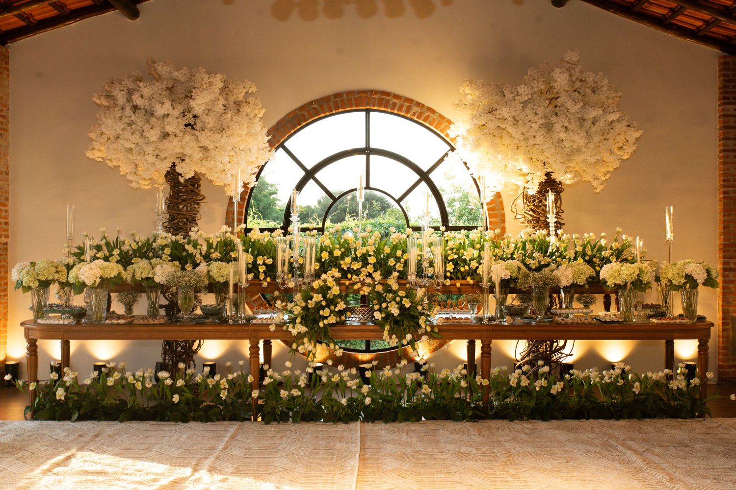 Elegant wedding reception floral display with white blooms, greenery, and candlelight against an arched window backdrop.