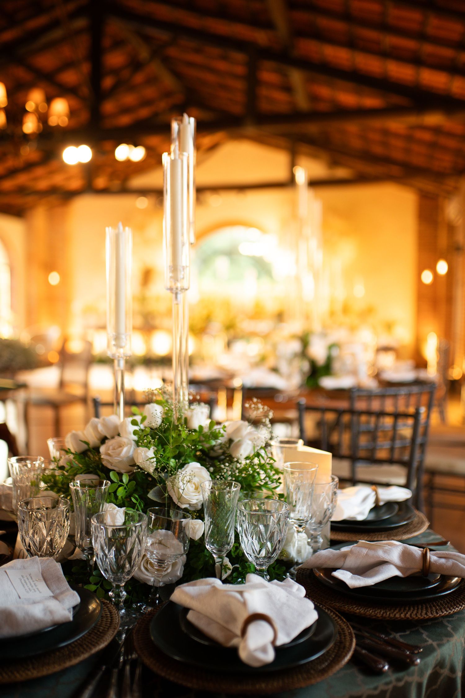 Elegant wedding reception table with white roses, tall candles, crystal glasses, and dark charger plates in a warmly lit venue.