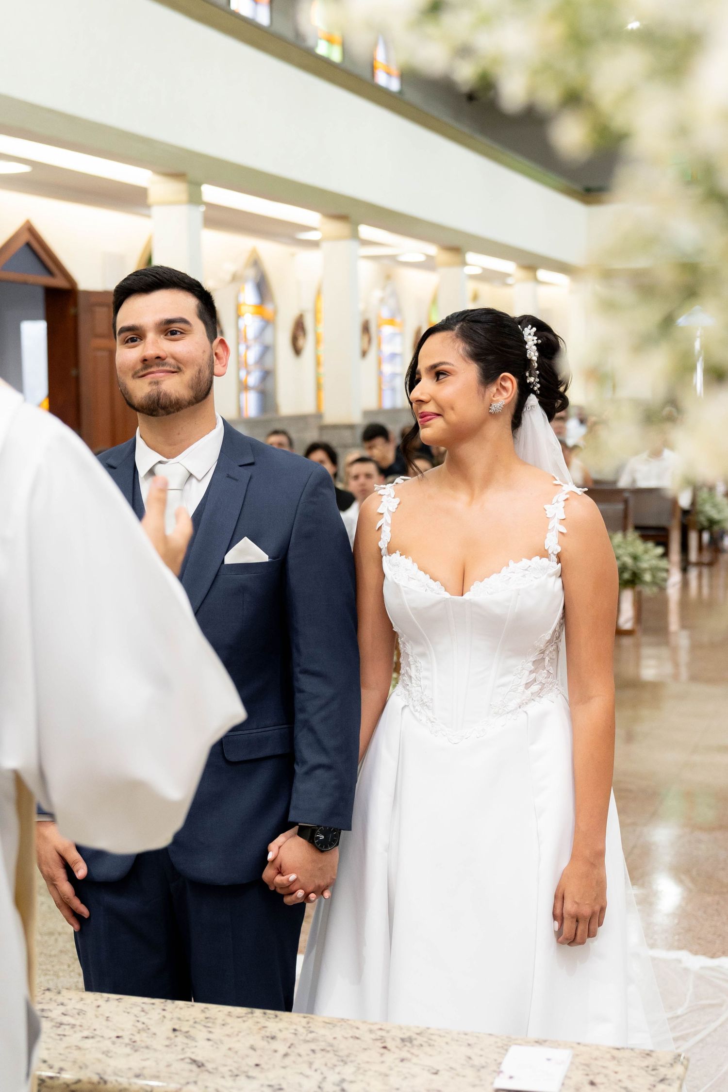 Bride in white lace gown and groom in navy suit hold hands at church altar during wedding ceremony.