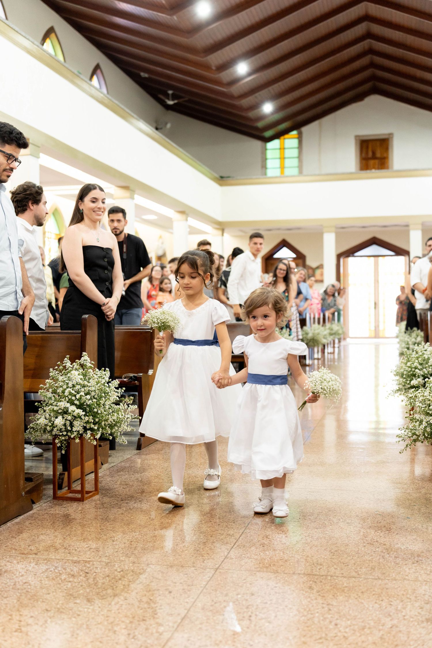 Two young flower girls in white dresses with blue sashes walk down a church aisle during a wedding ceremony.